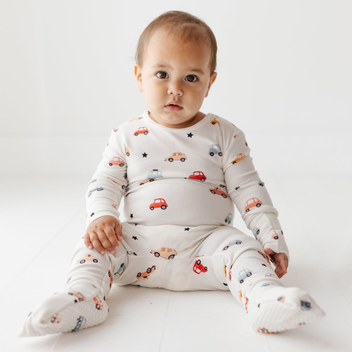 A baby sits on a white floor, wearing a white romper with a car-themed print.