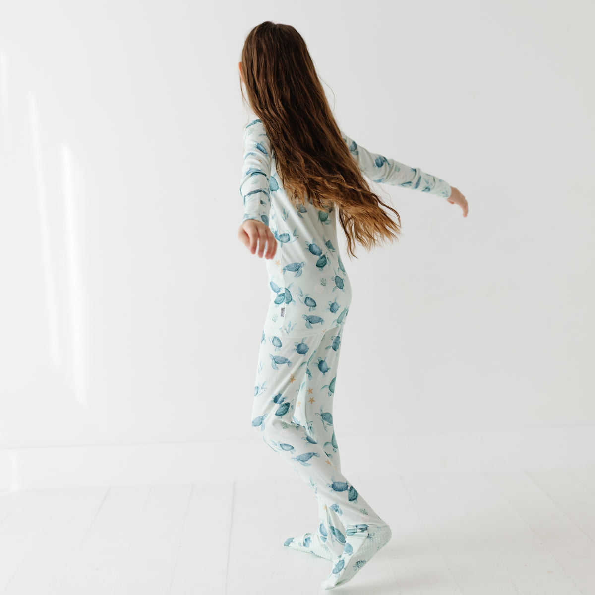A kid stands on a white floor wearing a light green pajama set with turtles on it.