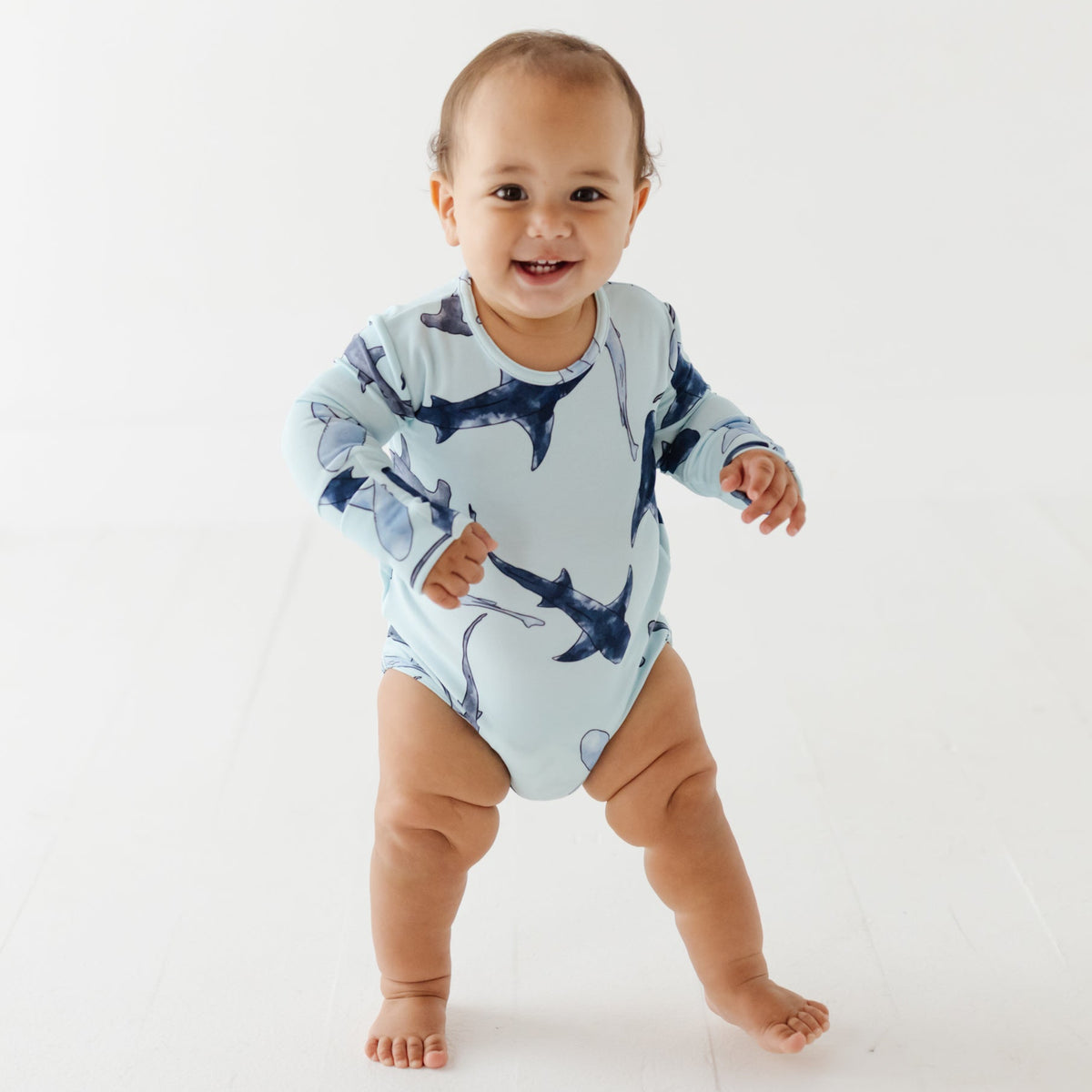 A smiling baby stand on white floor wearing a light blue onesie with different shades of blue sharks on it.