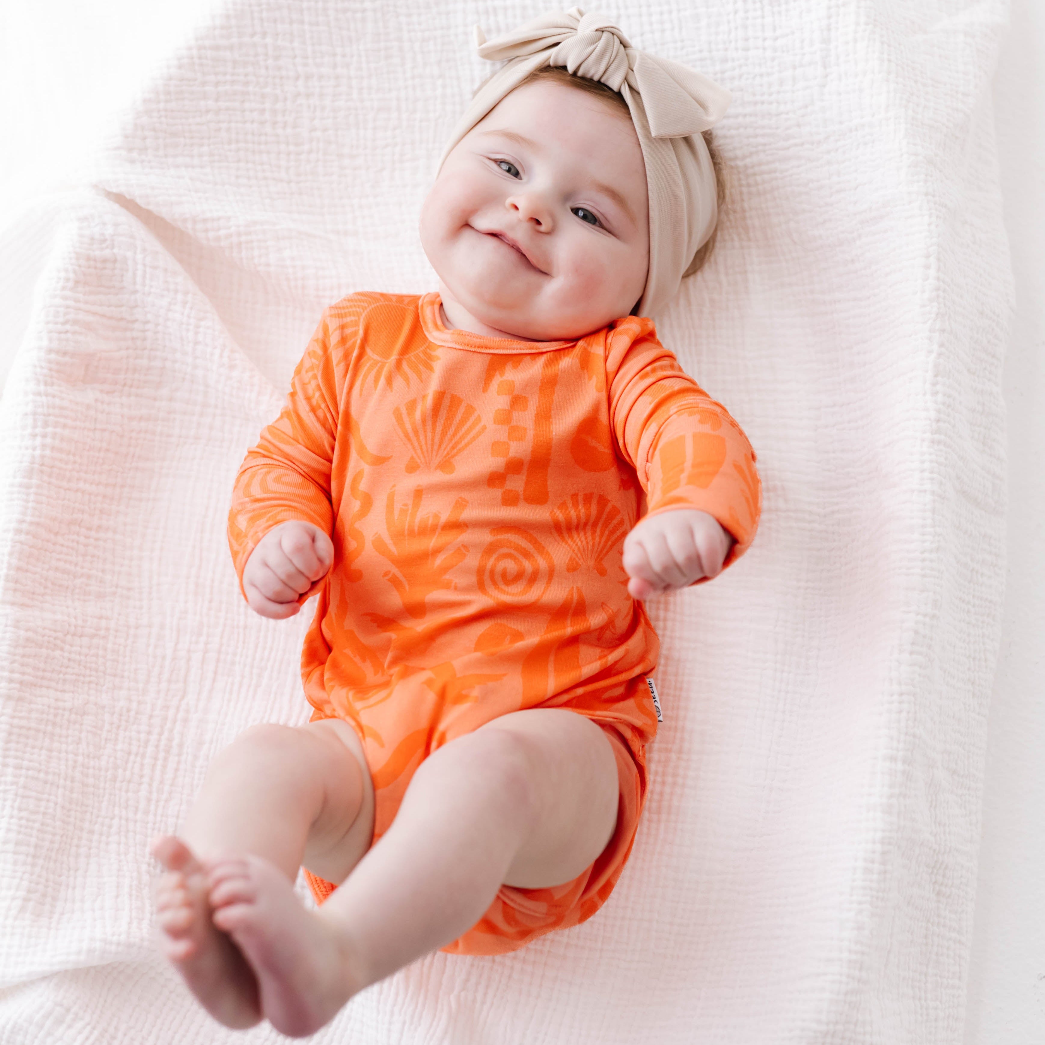 Baby in an orange onesie and beige headband lying on a white blanket