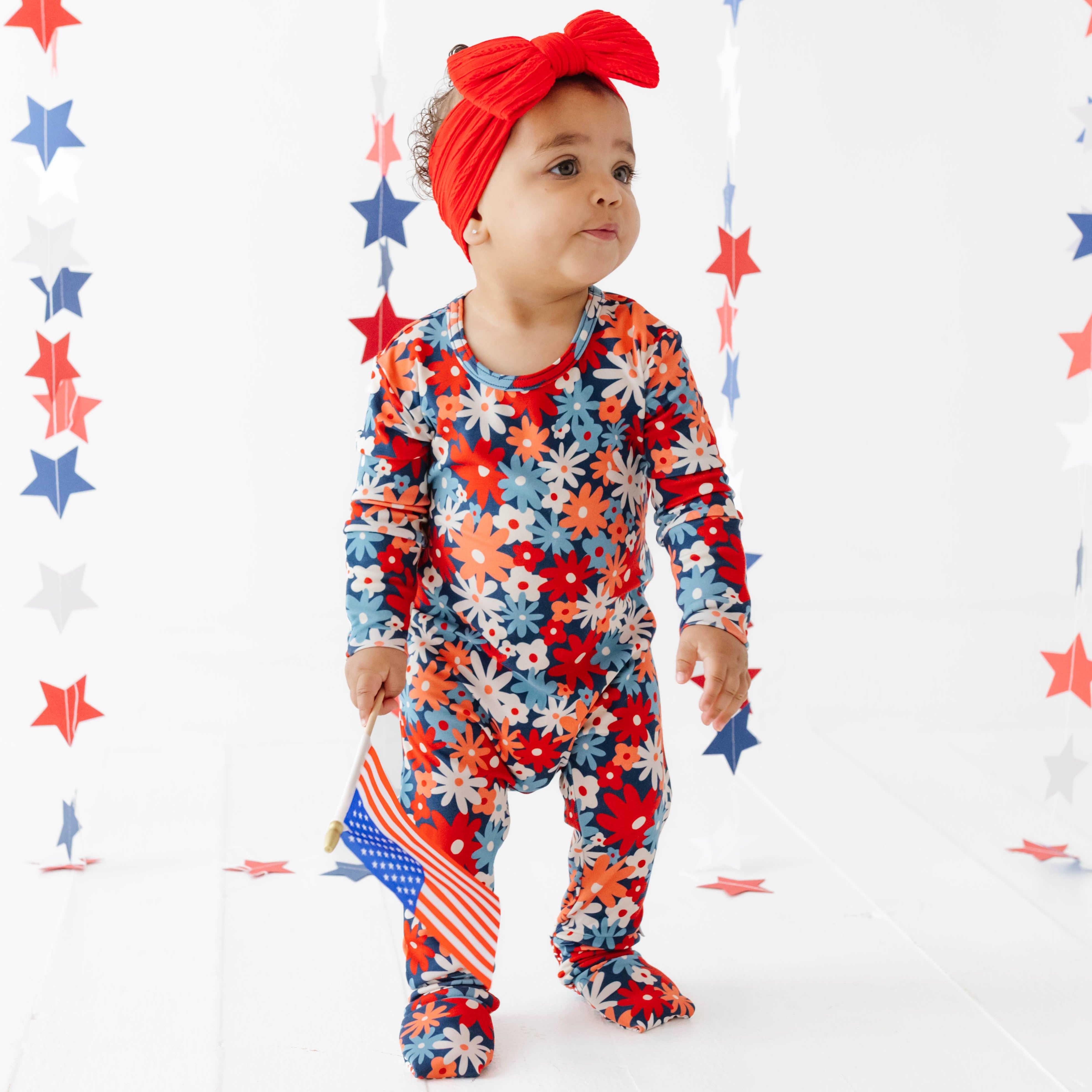 Baby in a colorful outfit with an American flag design, standing against a white background with red, blue, and gray stars.