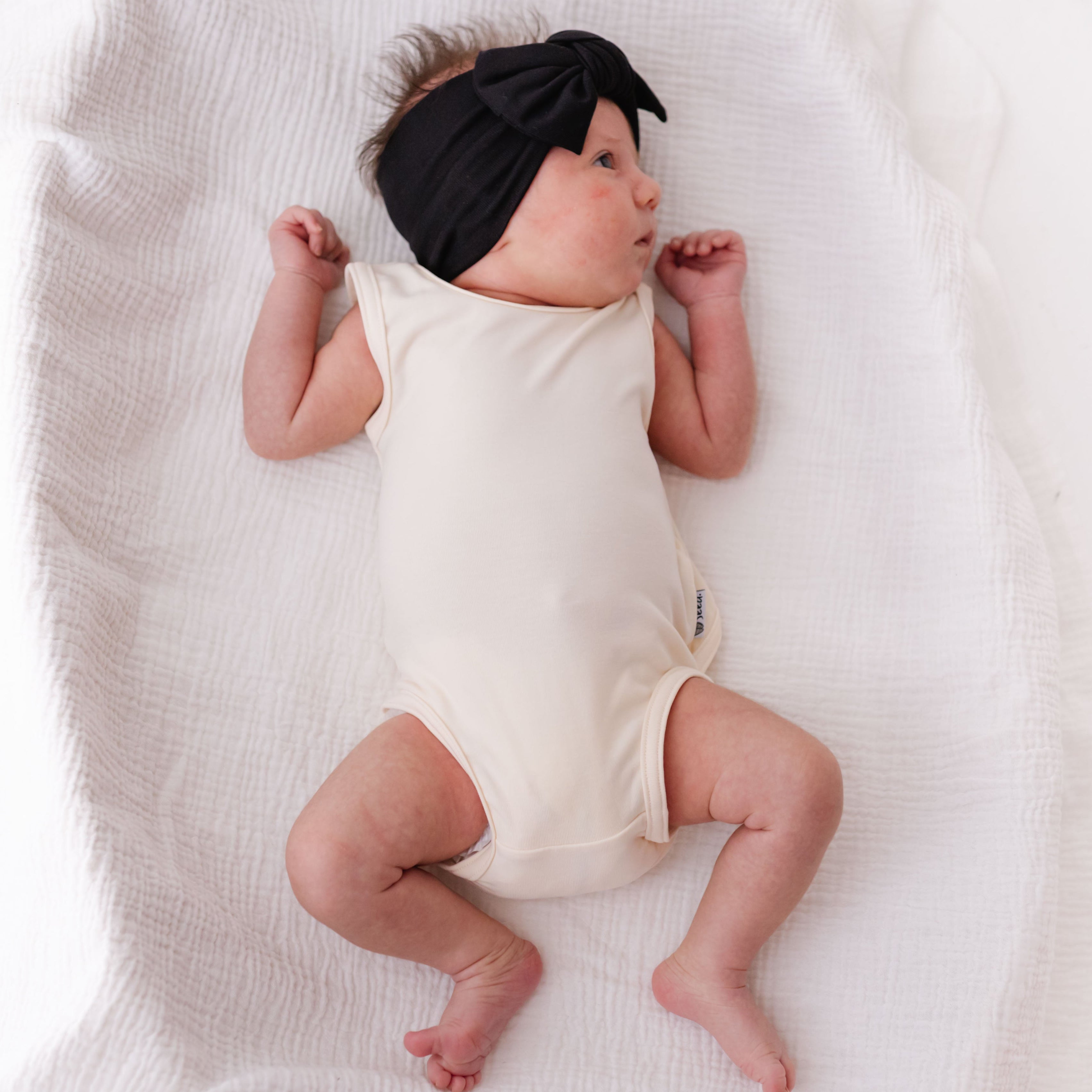 Baby lying on a white blanket wearing a black headband and cream-colored onesie.