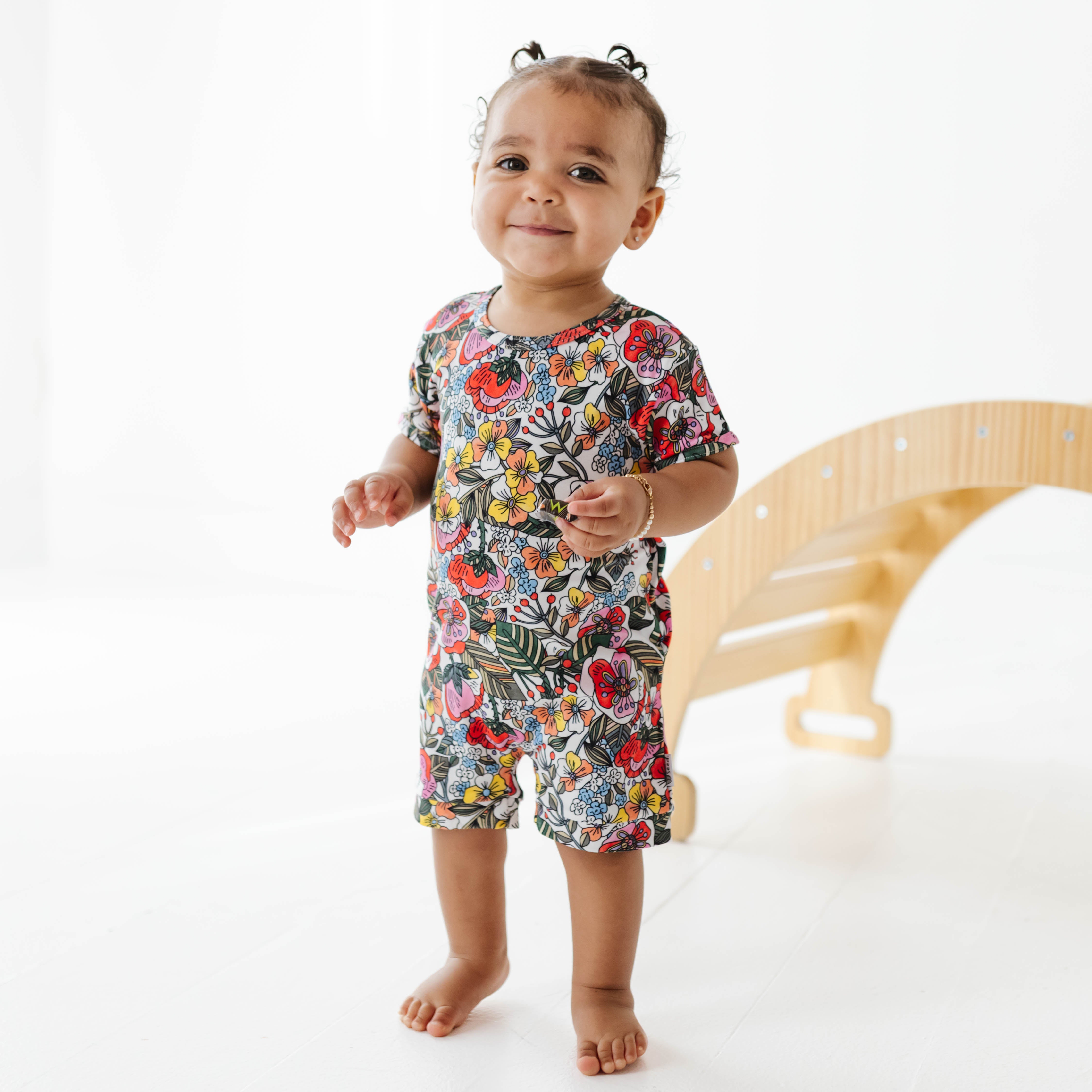 Child wearing a colorful floral romper standing on a white floor with a wooden toy in the background.