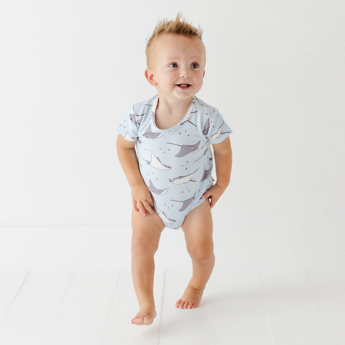 A baby stands on a white floor wearing a light blue onesie with a stingray pattern