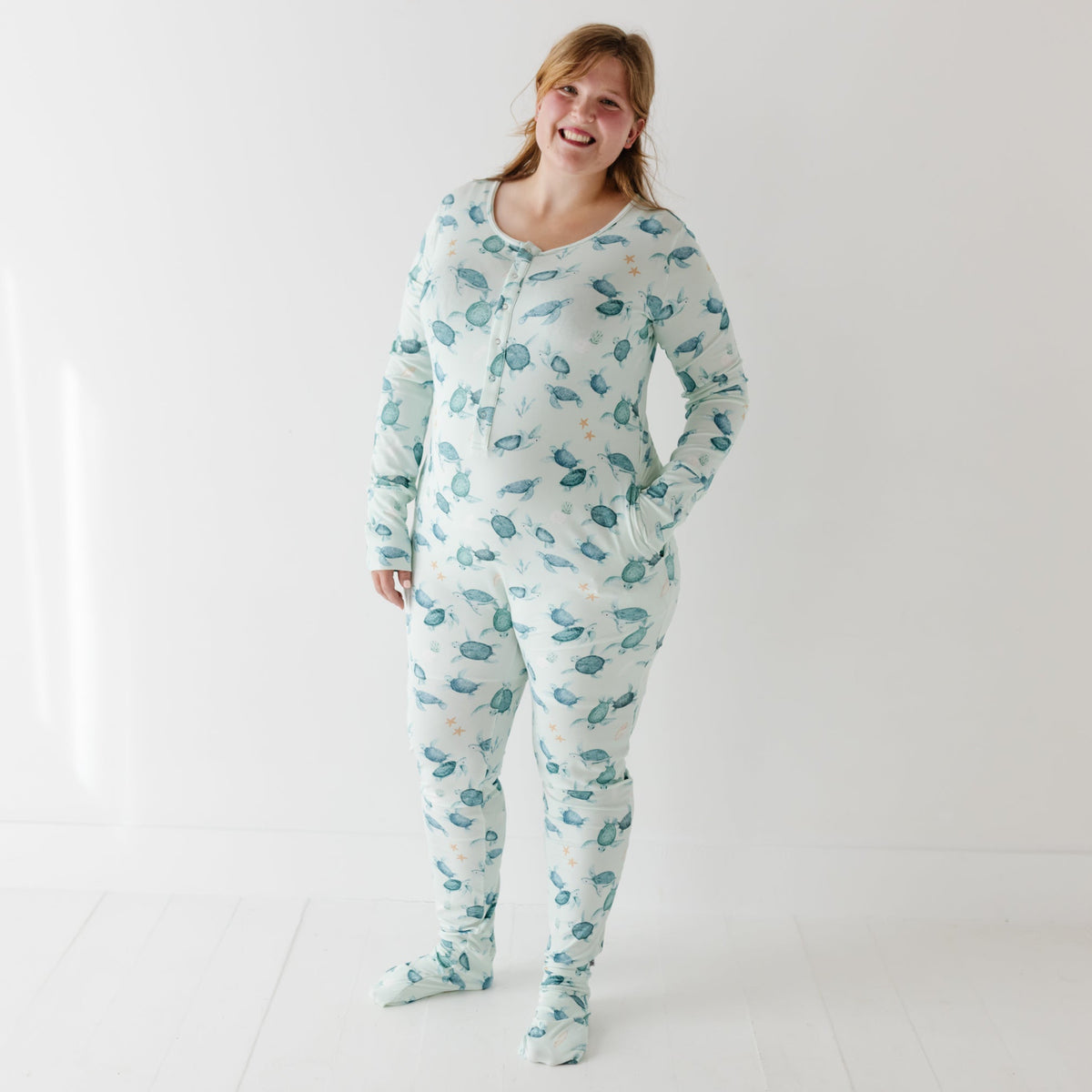 A smiling woman stand on white floor wearing a light turquoise romper with turtles on it.