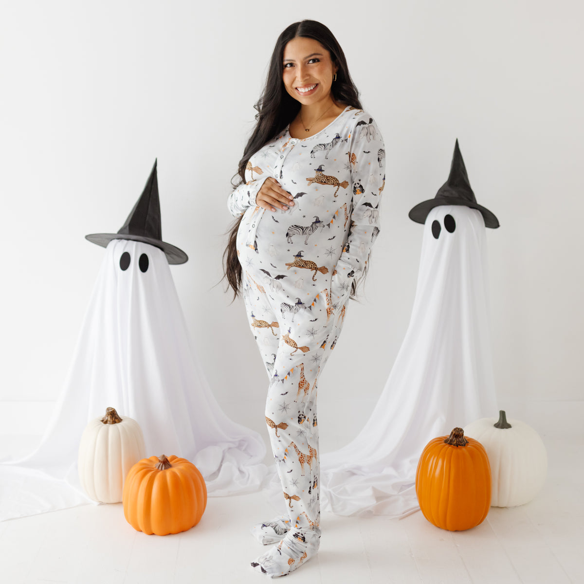 A smiling woman stand on white floor with ghost and pumpkins in the background. She is wearing a white romper with a Halloween-themed design.