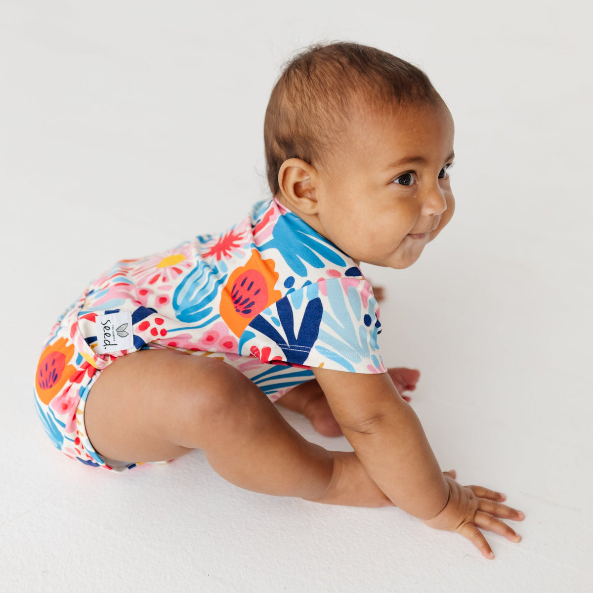 A smiling baby sit on a white floor wearing a colorful floral onesie.