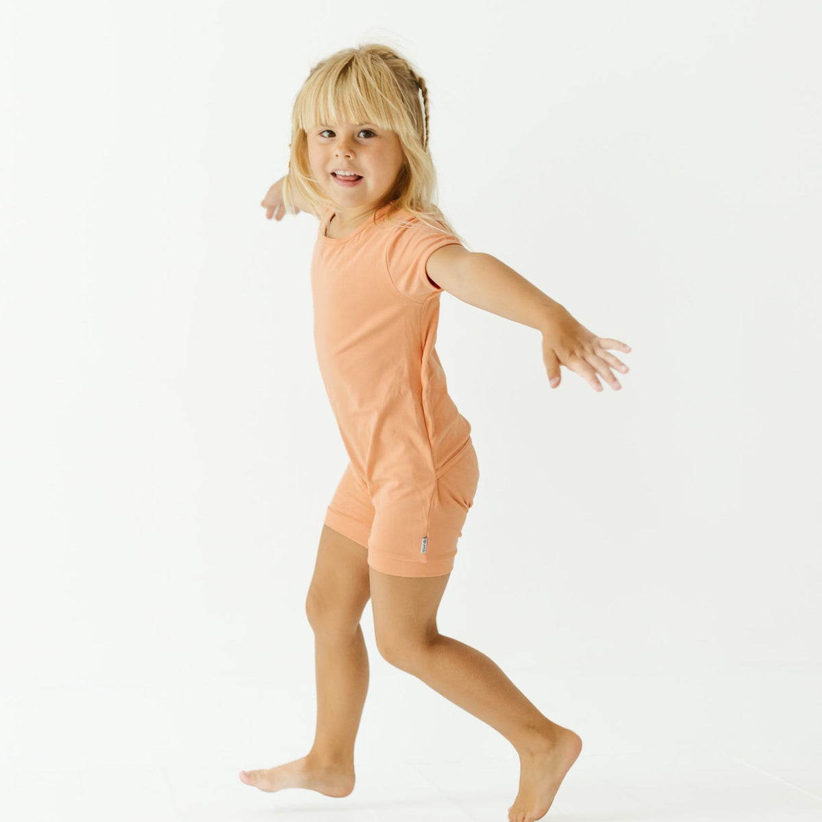 A young blonde girl wearing a peach-colored short-sleeve t-shirt and matching shorts, captured mid-step in a playful pose against a seamless white studio background.