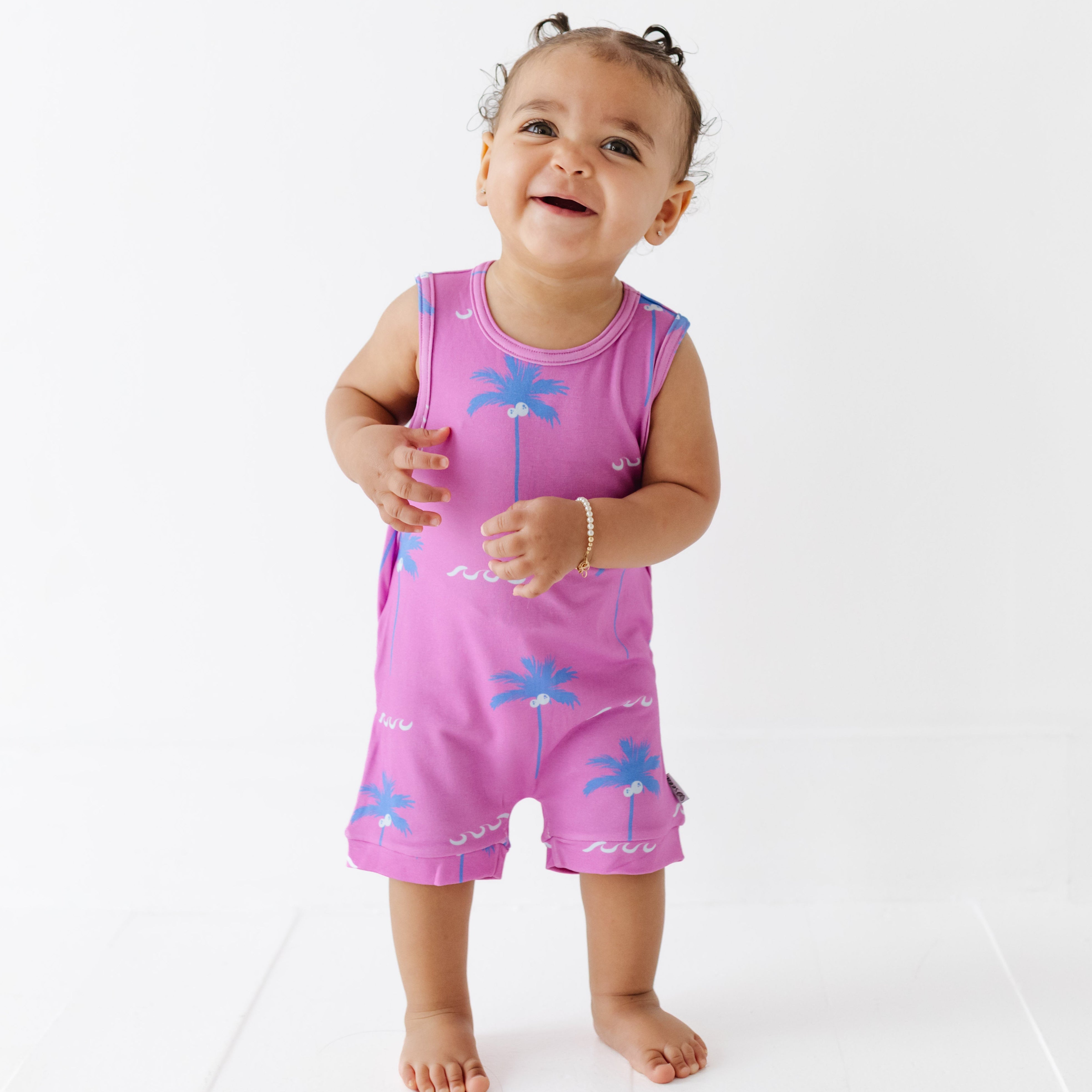 Baby wearing a pink sleeveless romper with blue palm tree designs on a white background