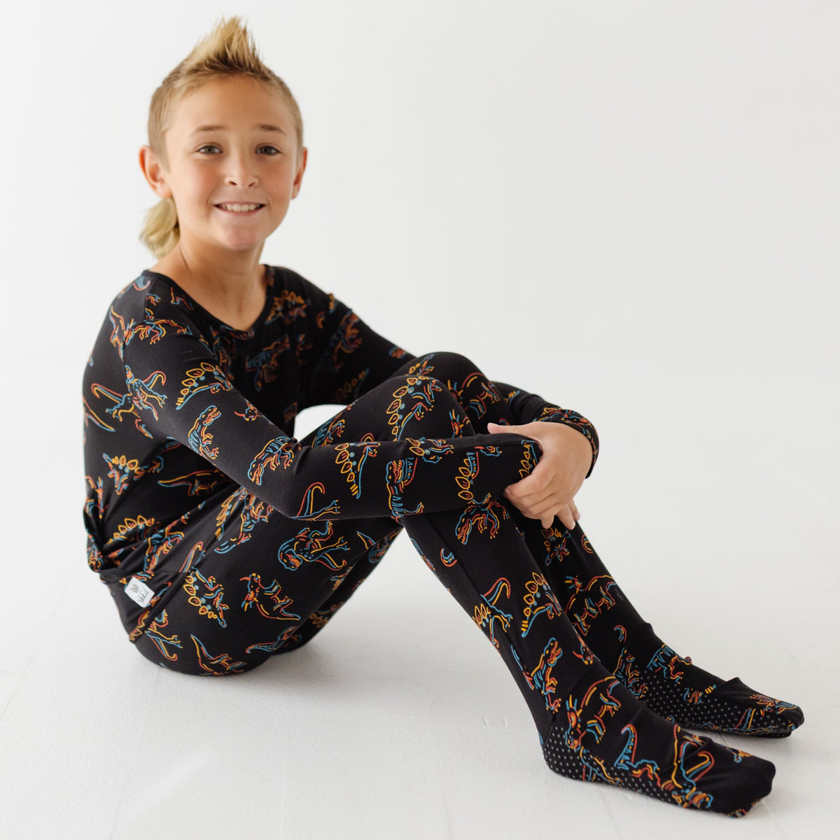 A kid sits on white floors wearing black romper with glowing dinosaurs on it.
