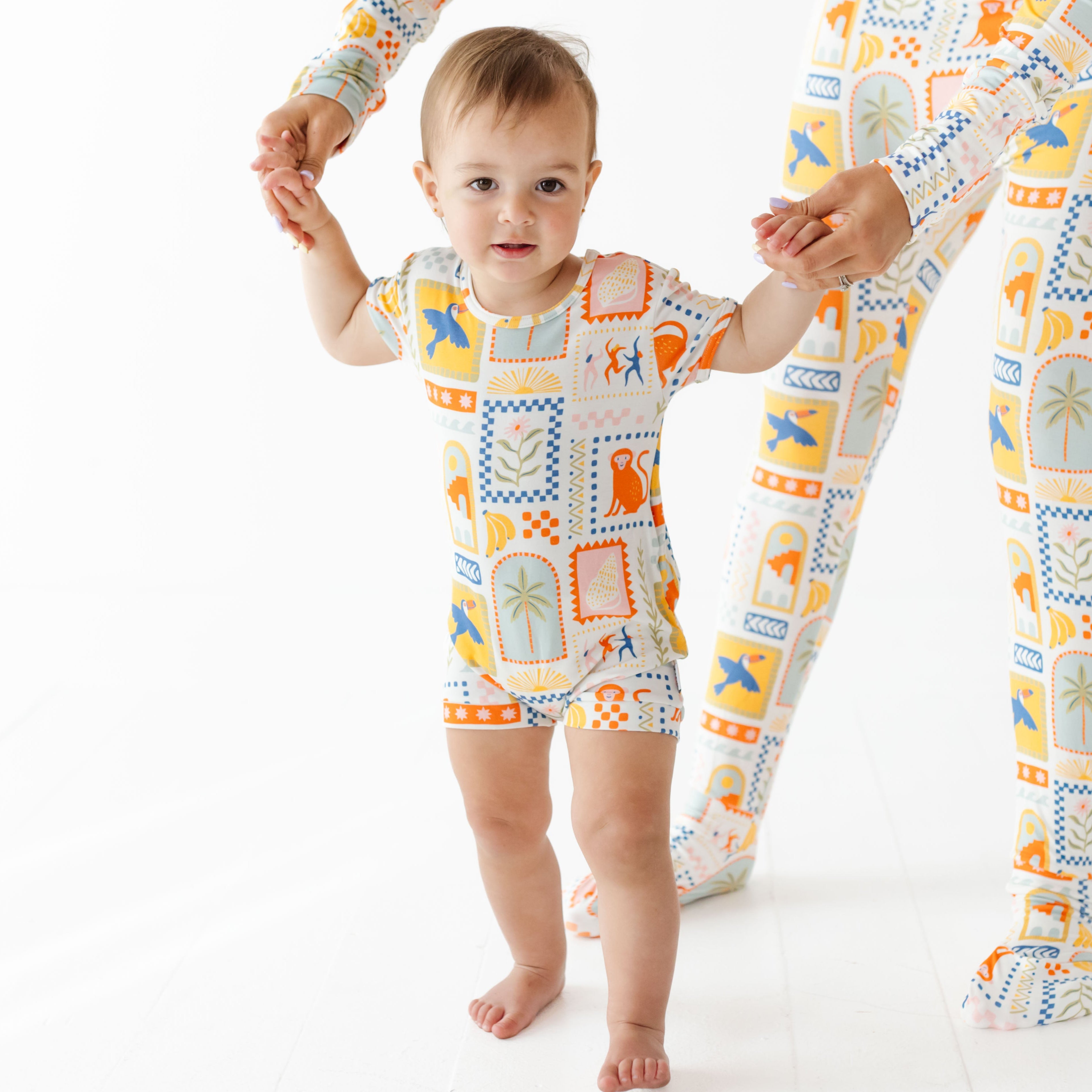 Baby in a colorful onesie standing next to a person wearing matching pajamas on a white background