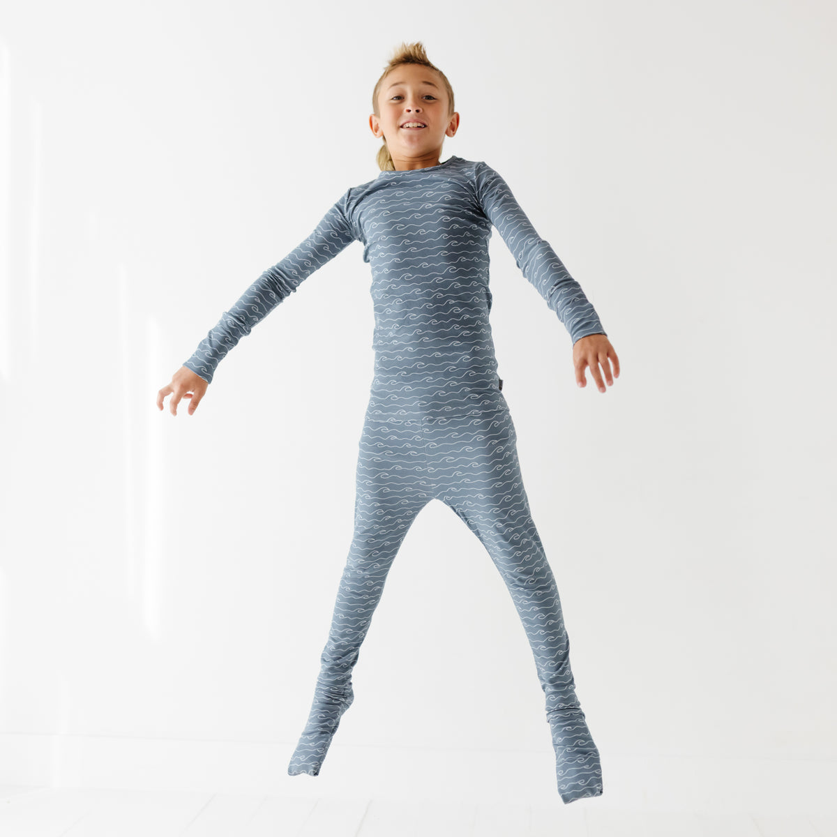 A smiling kid jumps on a white floor. They are wearing blue pajama set with waves in it.