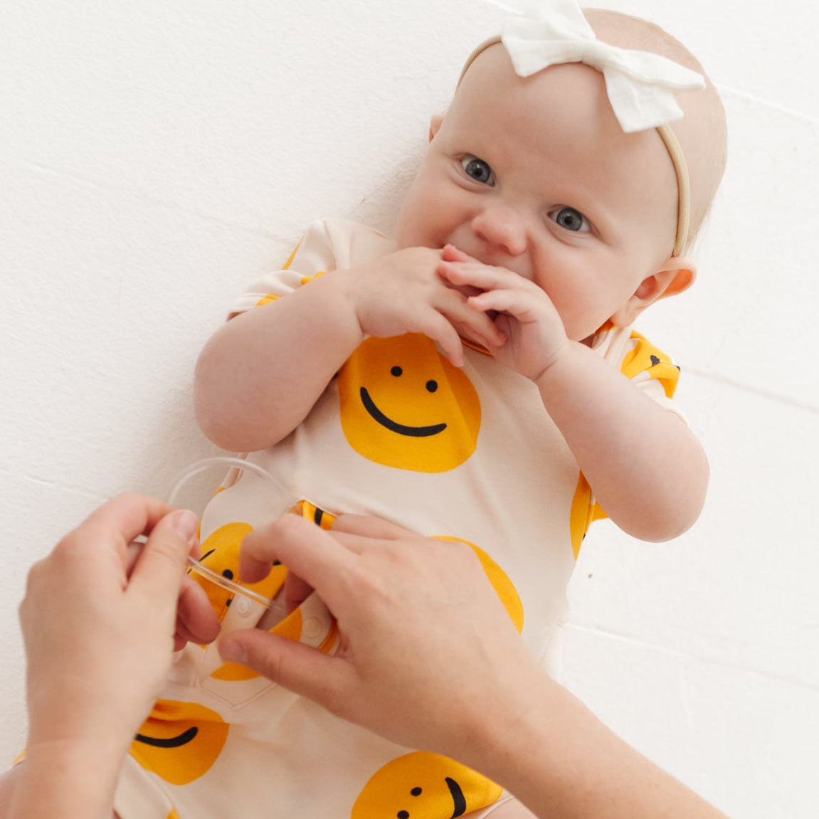 Baby wearing a onesie with smiley face patterns, sitting on a person's lap against a white background