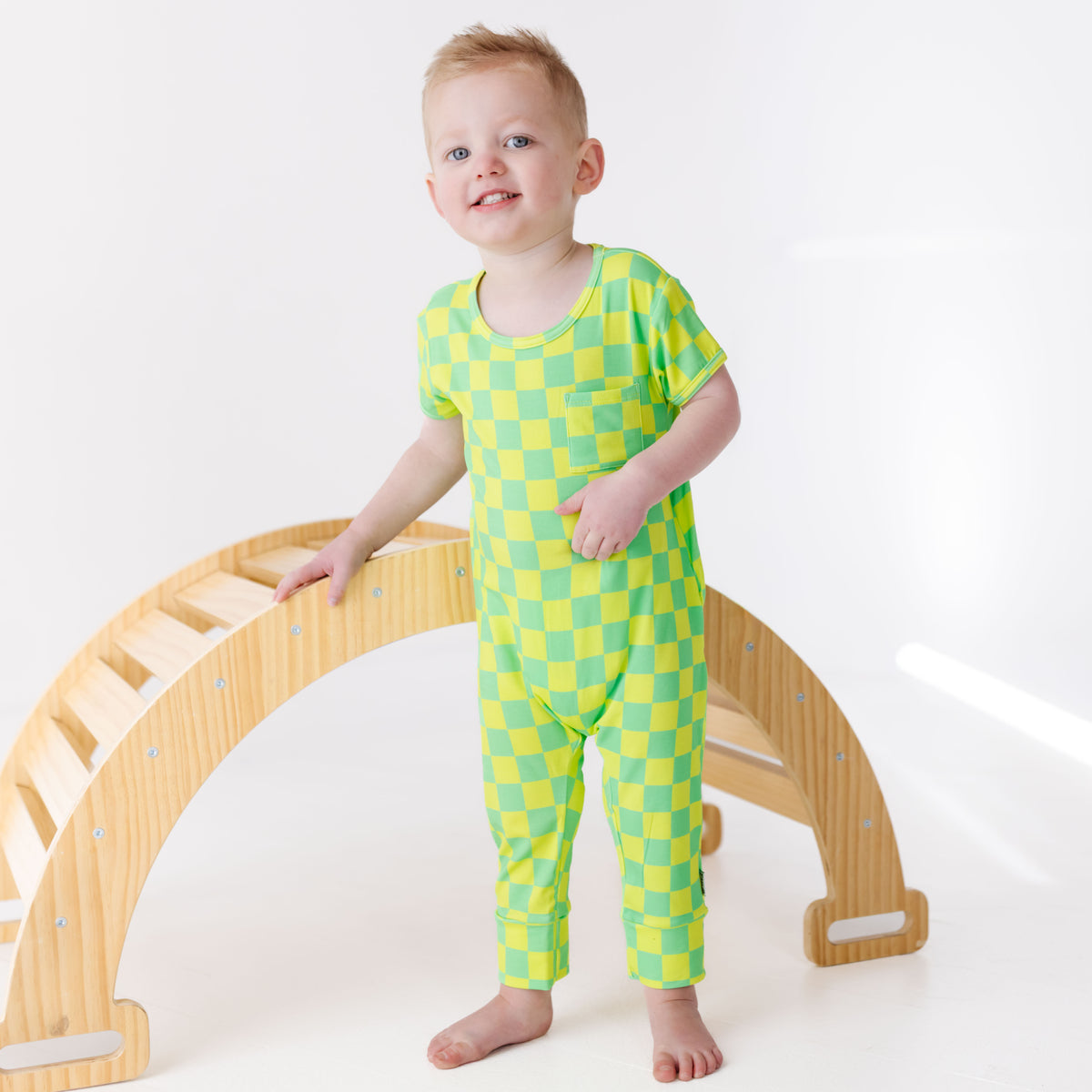 Child wearing a green and yellow checkered outfit standing next to a wooden arch toy on a white background