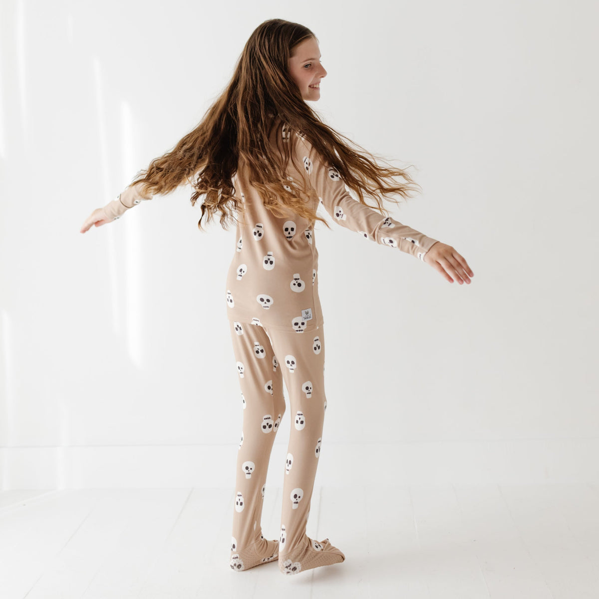 A smiling girl stands with her arms out on a white floor. She is wearing a tan pajama set with skulls on it.
