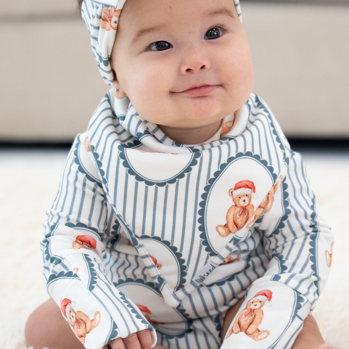 A baby sitting on a white shag rug wearing a matching long-sleeve romper and bow headband set with a blue and white pinstripe pattern and cartoon monkey illustrations.