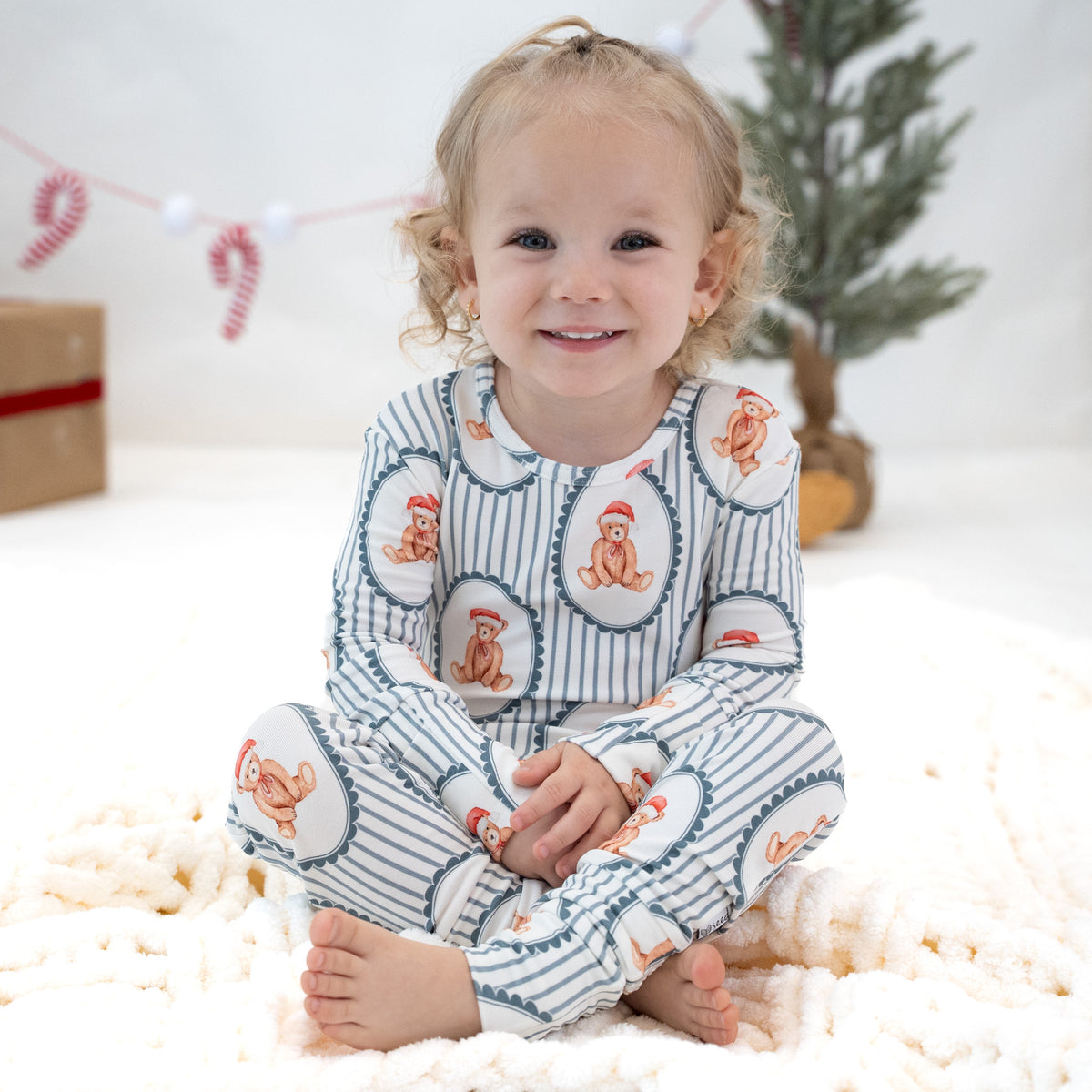 A smiling toddler sits cross-legged on a fluffy cream blanket, wearing a long-sleeve pajama set with blue and white vertical stripes and orange teddy bear motifs inside white hearts. The background is a soft, warm holiday setting with a small tree and candy cane garland.