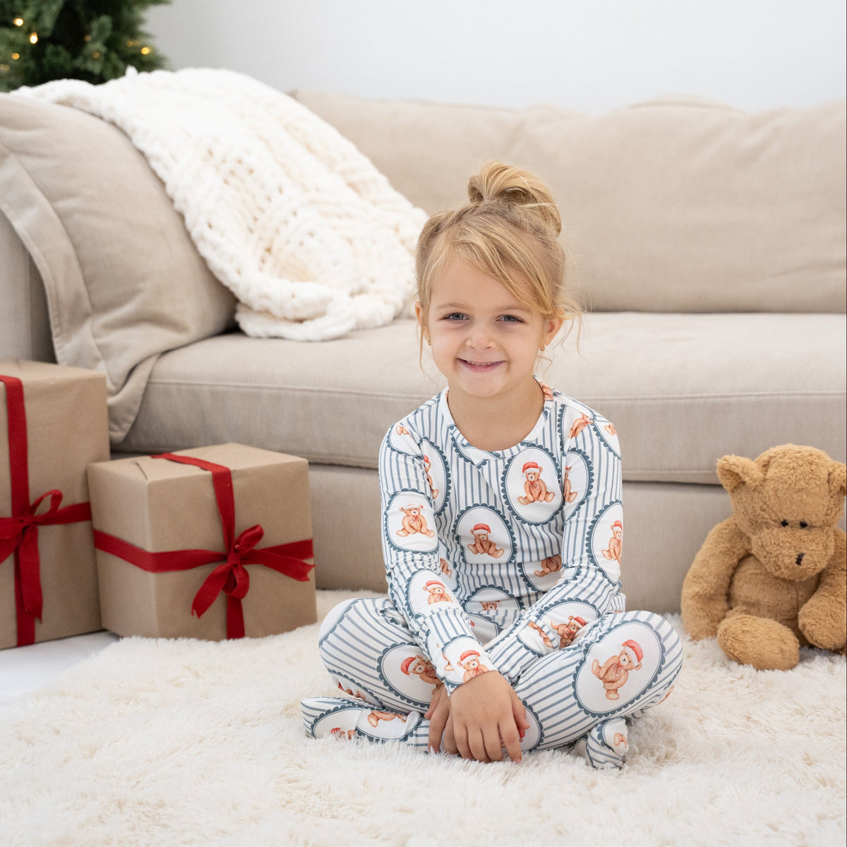 A young girl sits cross-legged on a white fluffy rug in a living room, wearing a long-sleeve pajama set with a blue and white striped pattern and cartoon teddy bear motifs. Two wrapped Christmas presents and a brown teddy bear are visible beside her, with a beige couch in the background.