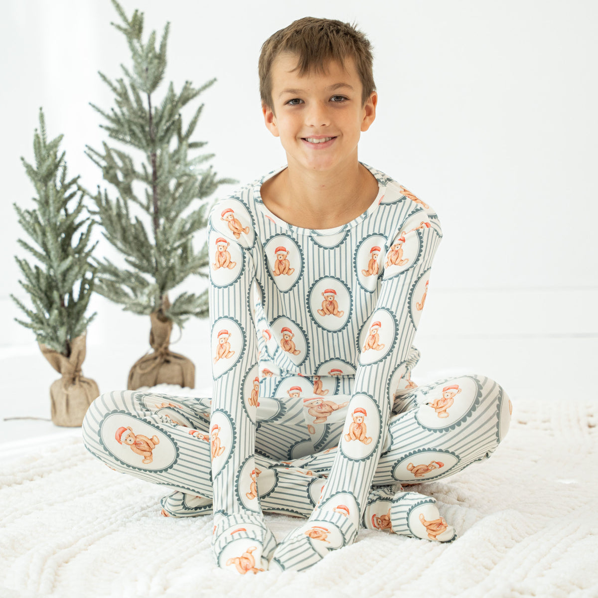 A smiling boy sits cross-legged on a white textured blanket wearing a matching long-sleeve pajama set with a striped and oval pattern. Two small artificial trees stand in the background against a white wall.