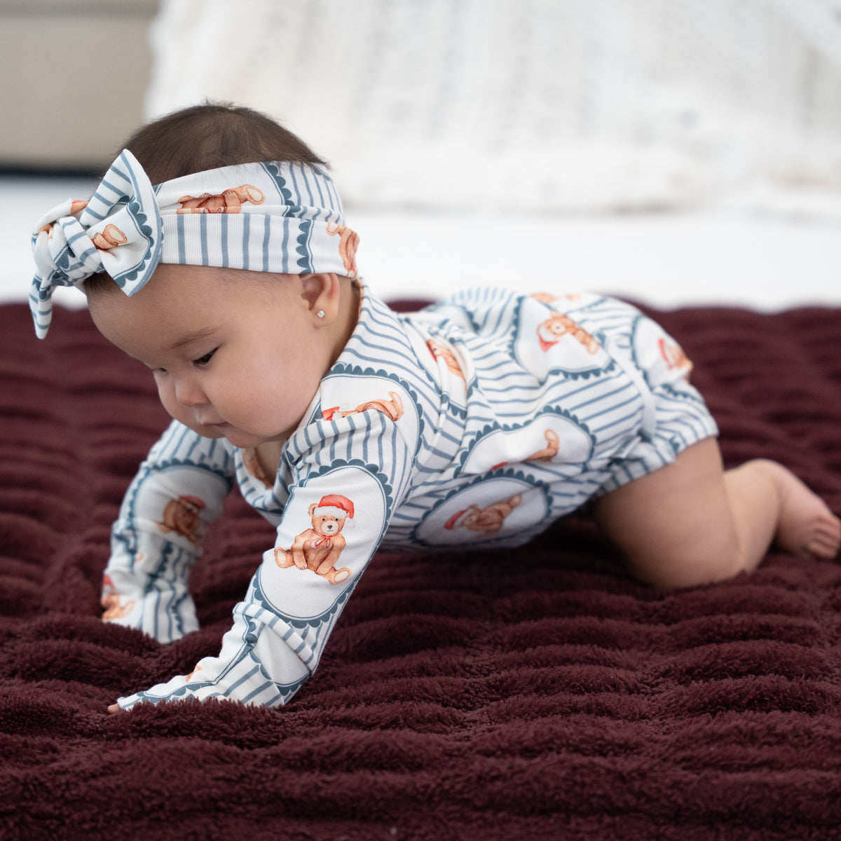 A baby crawling on a dark burgundy blanket, wearing a white and navy striped one-piece outfit with teddy bear illustrations and a matching striped bow headband.