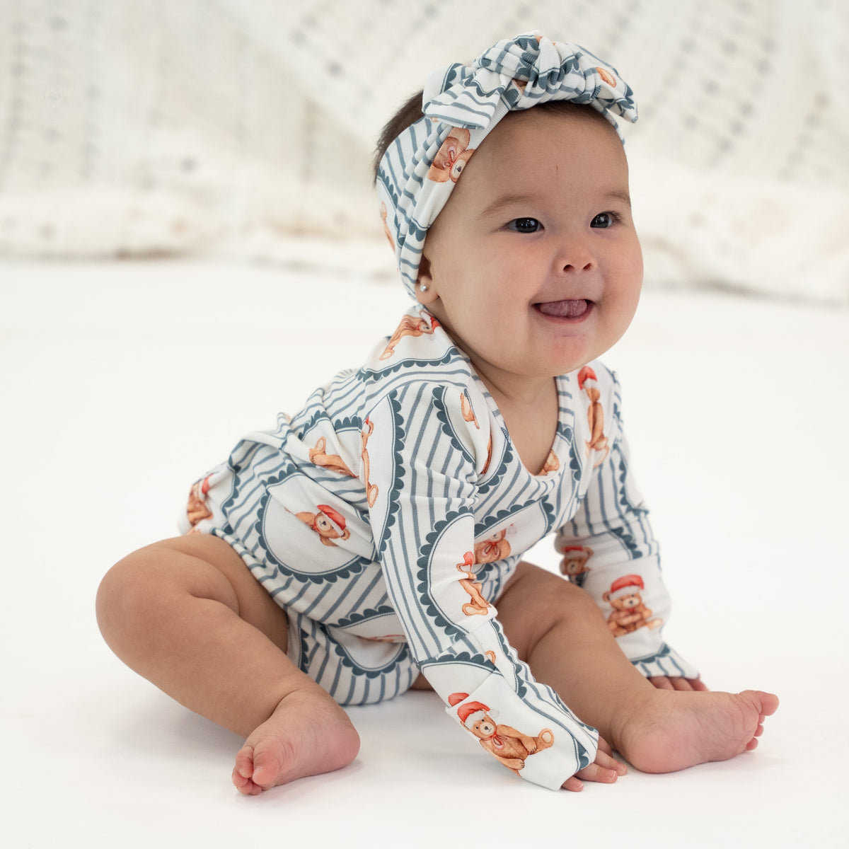 A baby sitting on a white floor wearing a matching long-sleeve romper and headband set with a blue and white striped pattern and orange cat illustrations.