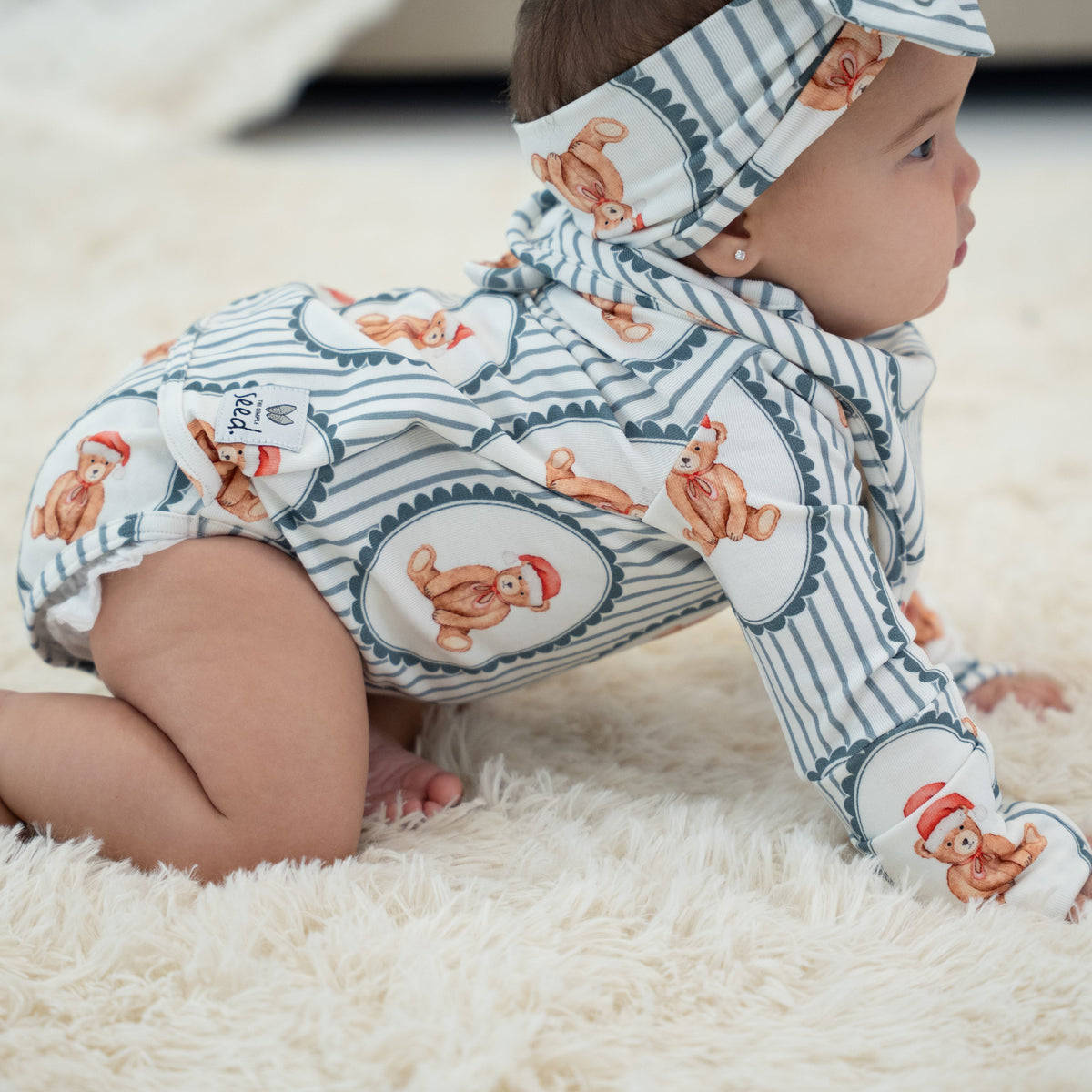 A baby crawling on a fluffy white rug, wearing a matching long-sleeve bodysuit and headband set with a white and navy blue stripe pattern and teddy bear prints.