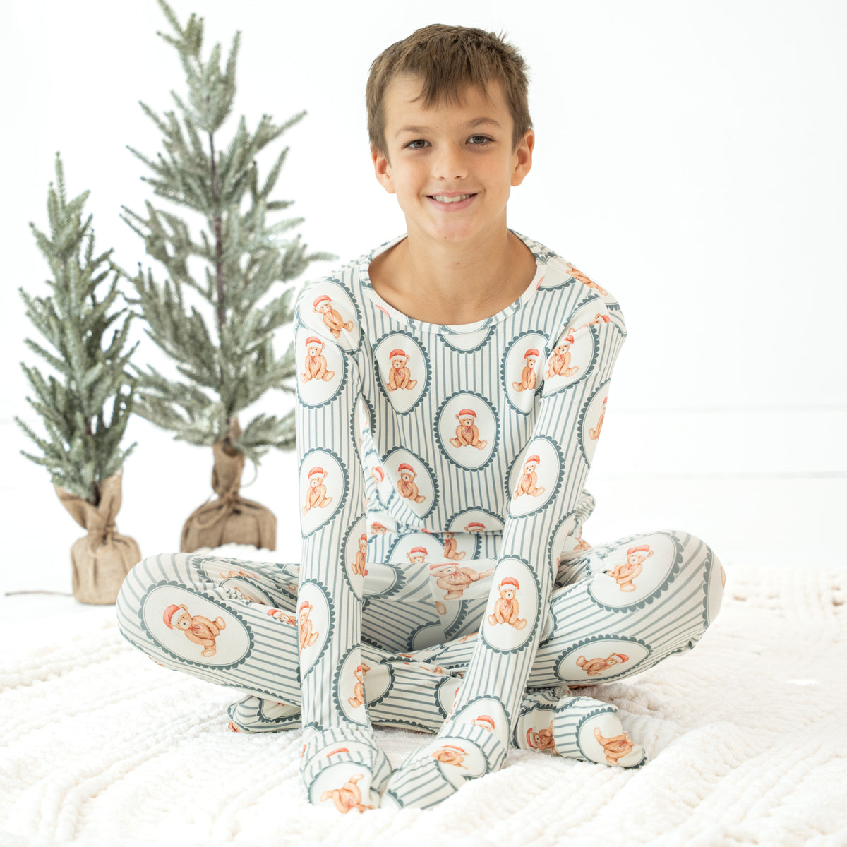 A young boy sitting cross-legged on a white blanket, wearing a matching long-sleeve pajama set with a striped pattern and orange cartoon monkey faces. Two small potted trees are in the background against a white wall.