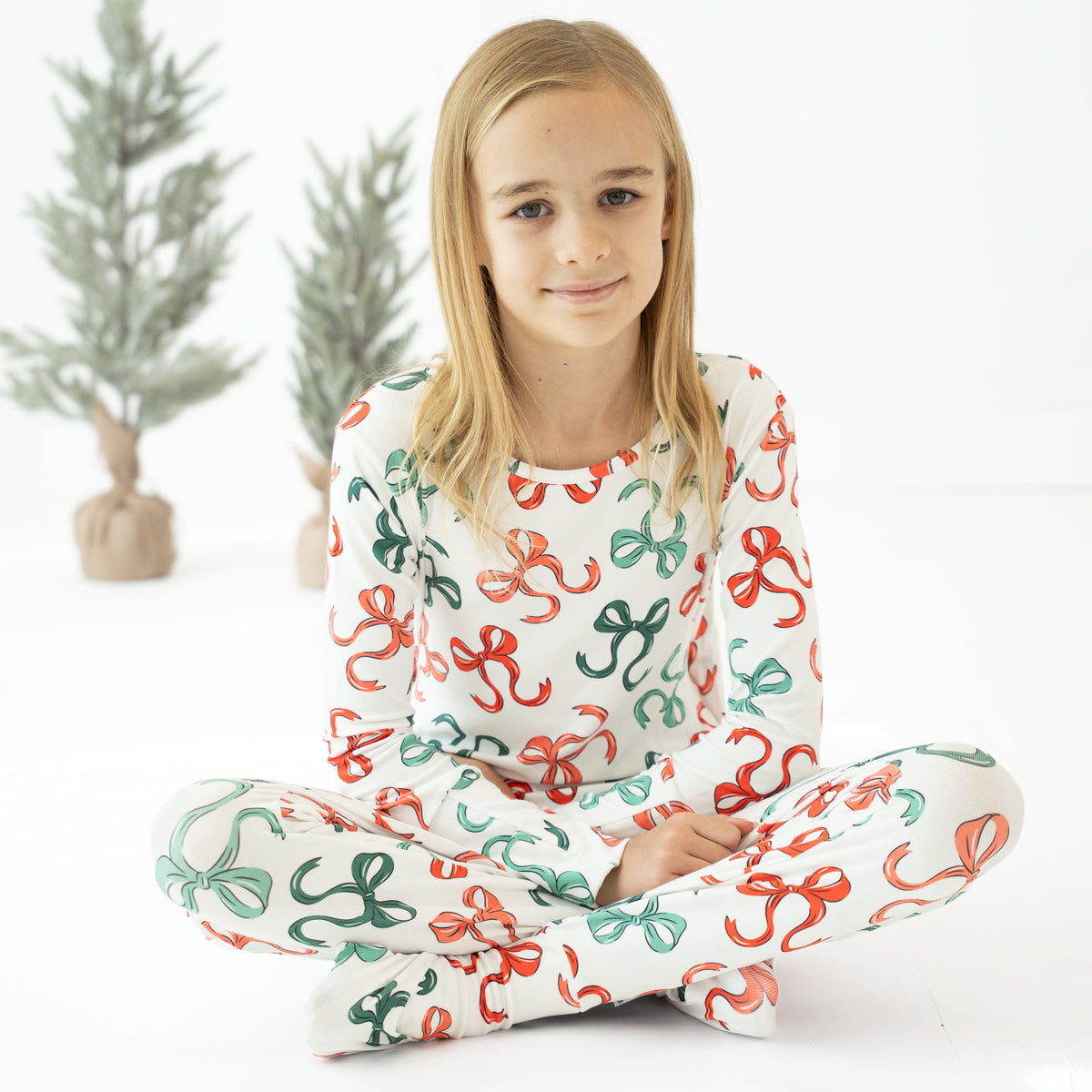 A young girl with blonde hair sits cross-legged on a white floor wearing a matching long-sleeve pajama set with a festive red and green bow pattern. Two small decorative Christmas trees are blurred in the background.