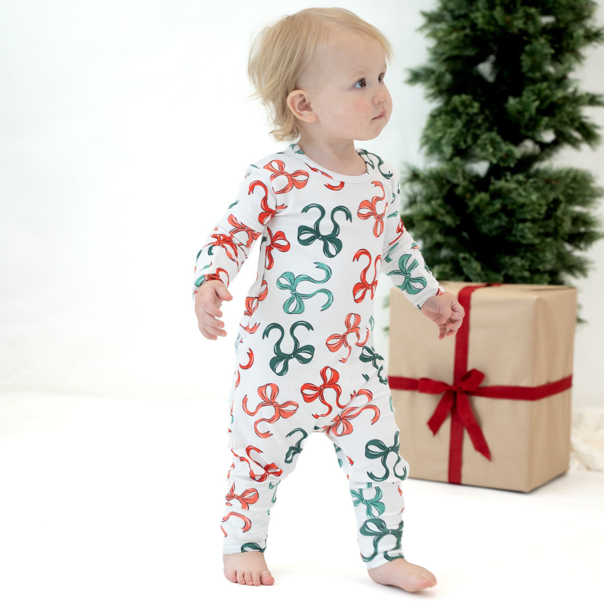 A baby wearing a white footed sleeper with a red and green ribbon pattern, standing next to a wrapped gift box and a Christmas tree.