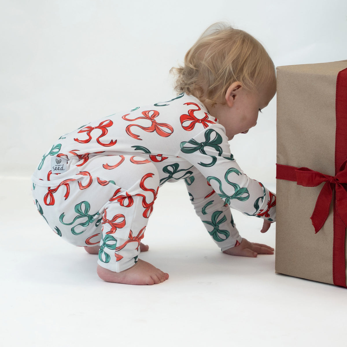 A baby wearing a white long-sleeve jumpsuit with a red and green ribbon pattern, crawling on a white studio floor next to a brown gift box with a red ribbon.