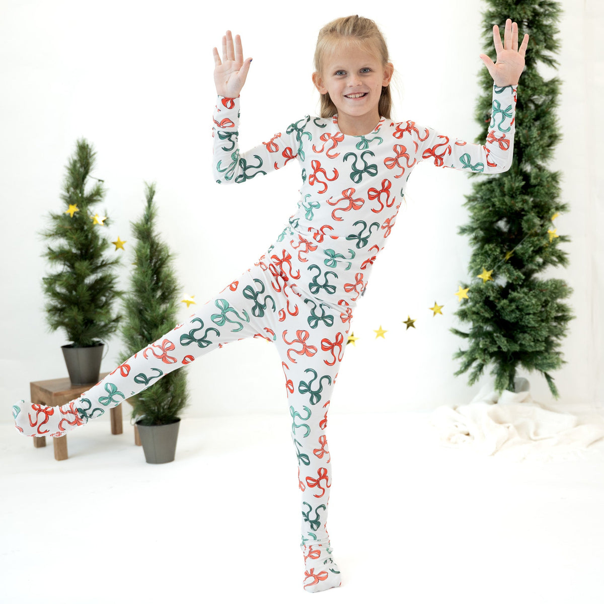 A young girl wearing a white full-body one-piece pajama or costume with a colorful repeating bow pattern, posing dynamically with one leg extended in a studio decorated with miniature Christmas trees and gold star accents.