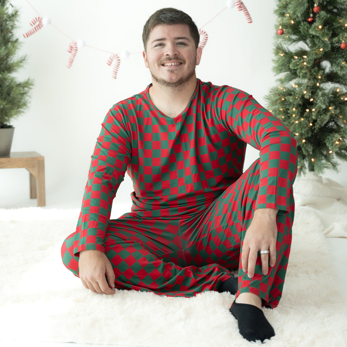 A man sitting on a white fluffy rug wearing a red and green checkered long-sleeve pajama set, with a decorated Christmas tree and candy cane garland in the background.