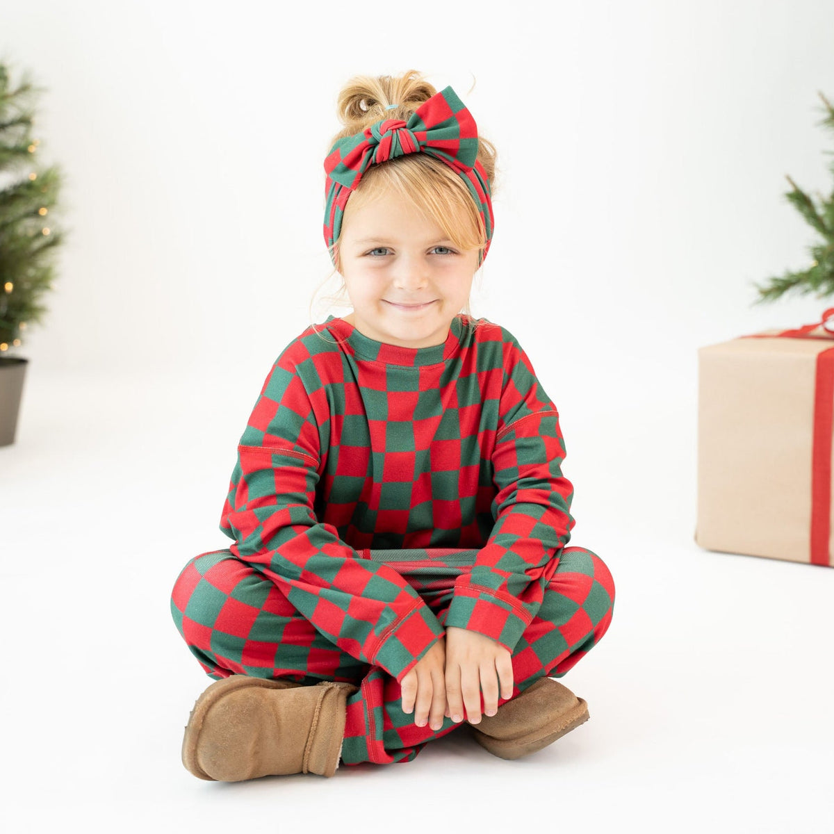 A young child sitting cross-legged on a white floor, wearing a red and green checkered long-sleeve pajama top and matching pants with a matching red and green checkered bow headband. The background is a clean white studio setting with Christmas trees and a wrapped gift box in the distance.