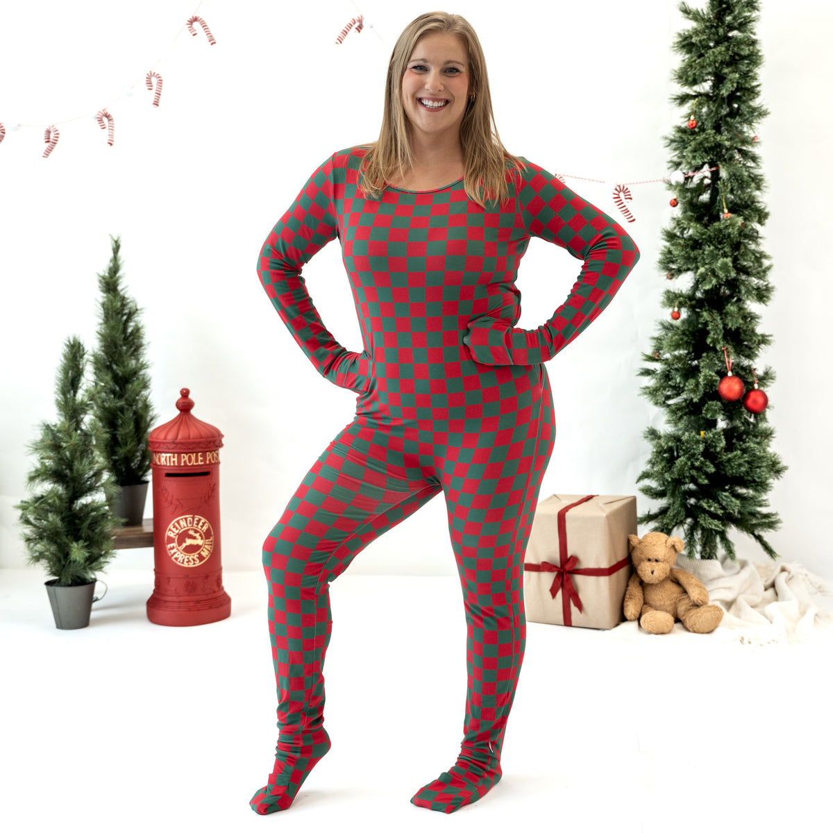 A woman modeling a red and green checkered holiday footie pajama against a white studio background decorated with Christmas trees, a red fire station prop, and a wrapped gift box.