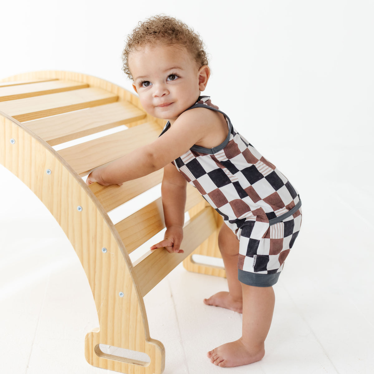 Child in a checkered outfit standing on a wooden step stool against a white background