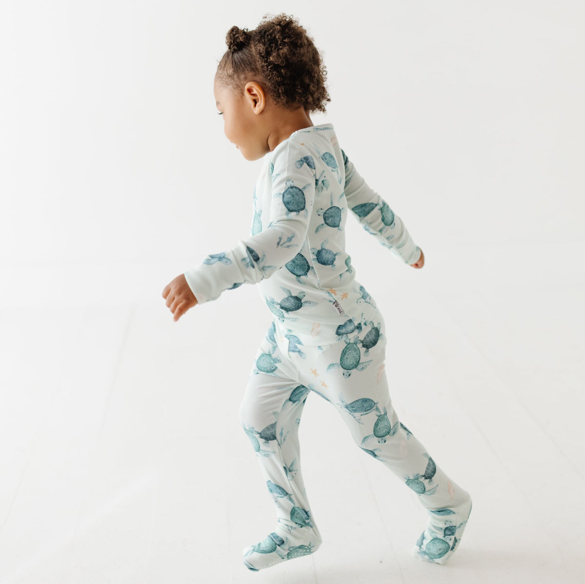 A kid stands on a white floor wearing a light green pajama set with turtles on it.