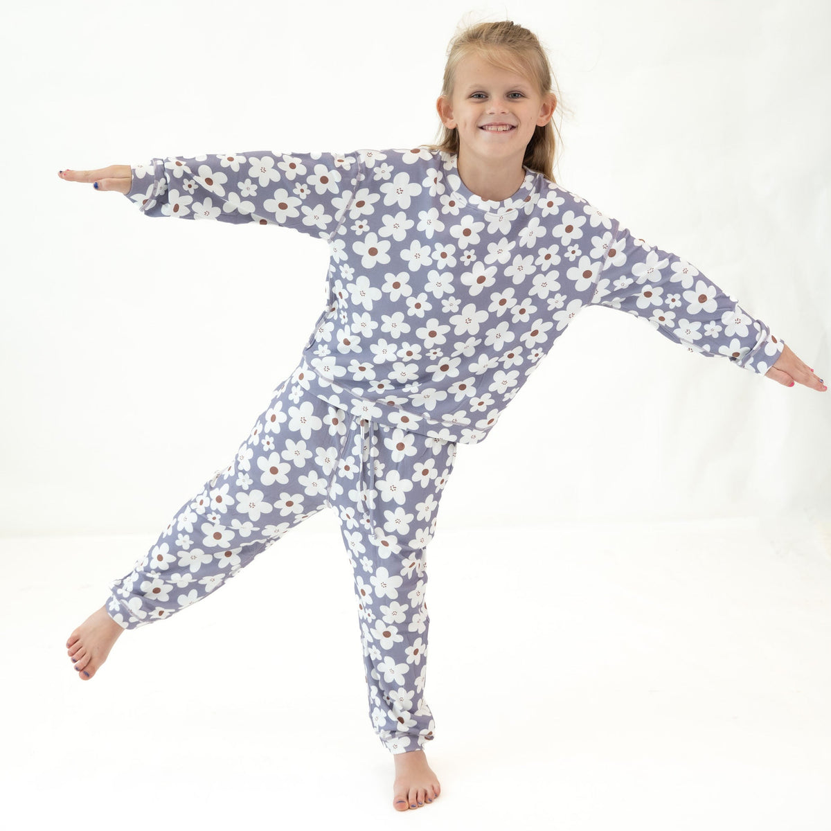 A young girl smiling and posing in a matching long-sleeve pajama set with a white daisy floral print on a dusty lavender-blue background, set against a plain white studio backdrop.