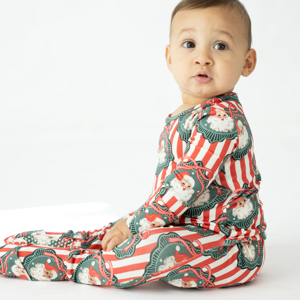 A baby sitting and wearing a festive Christmas print footed sleeper with red and white stripes and Santa Claus motifs, photographed against a clean white background.