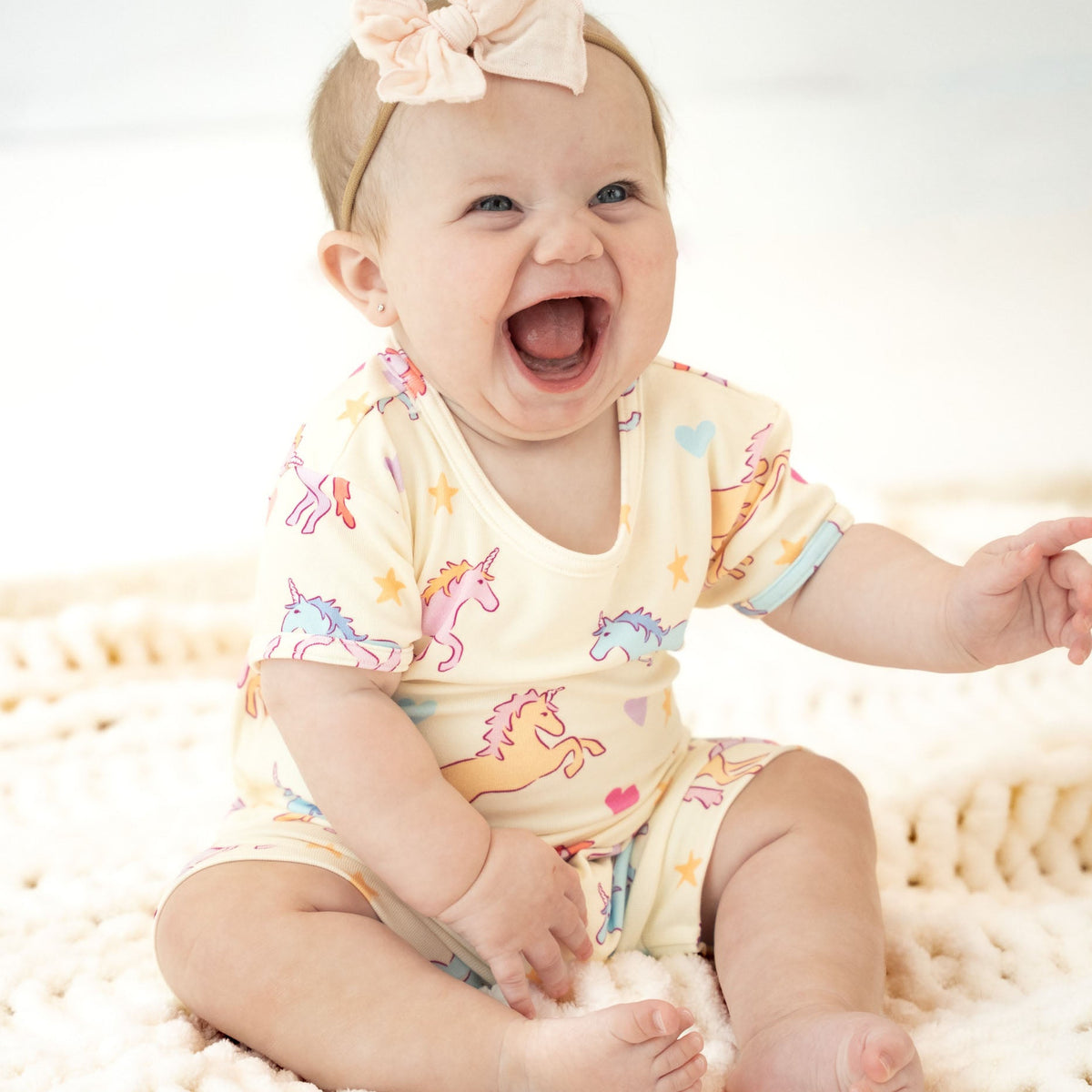 A baby girl sitting on a cream knit blanket, wearing a pale yellow short-sleeve romper with a colorful unicorn print and a matching pink bow headband.