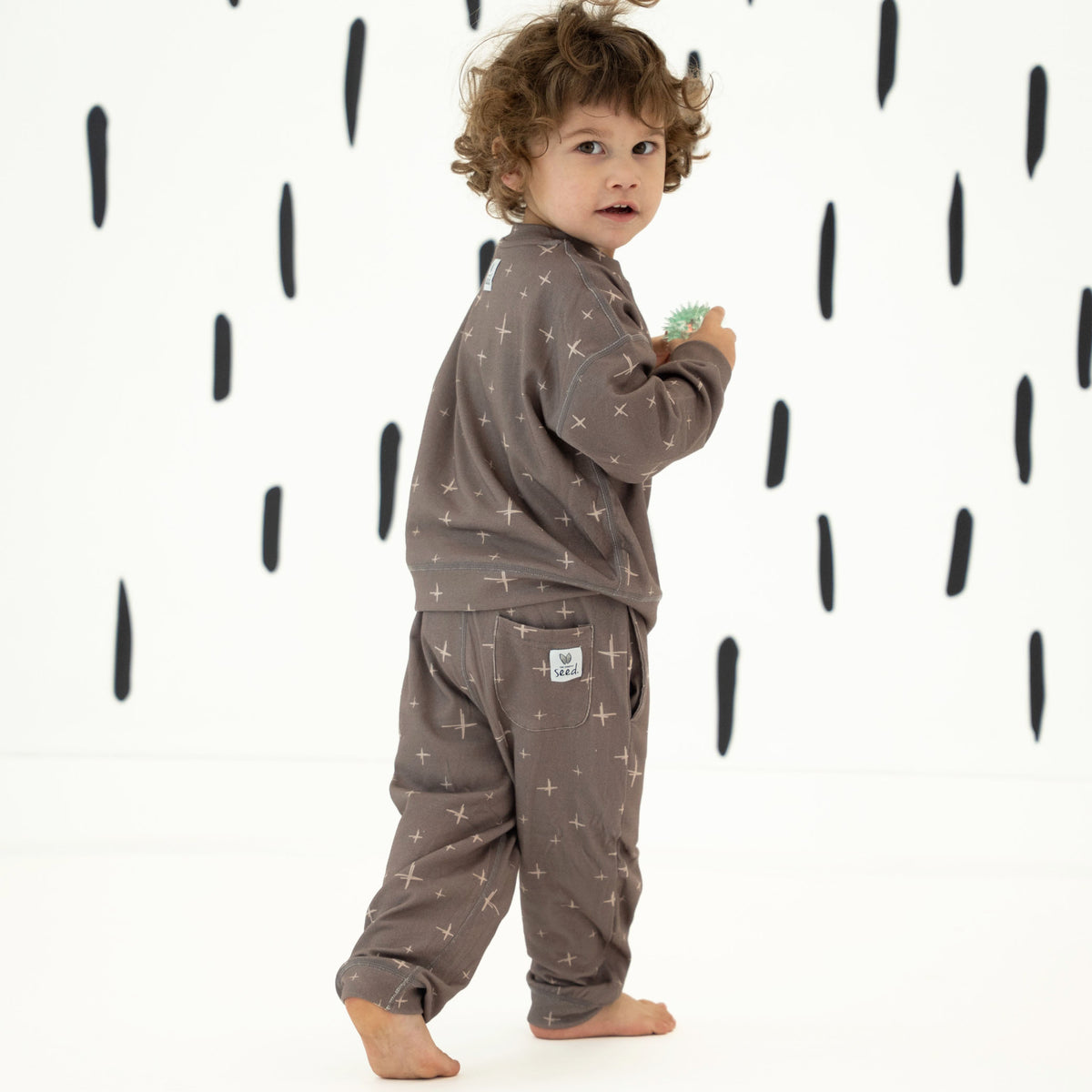 A toddler standing barefoot in a matching brown pajama set with a subtle star pattern, photographed against a white background with black raindrop shapes.