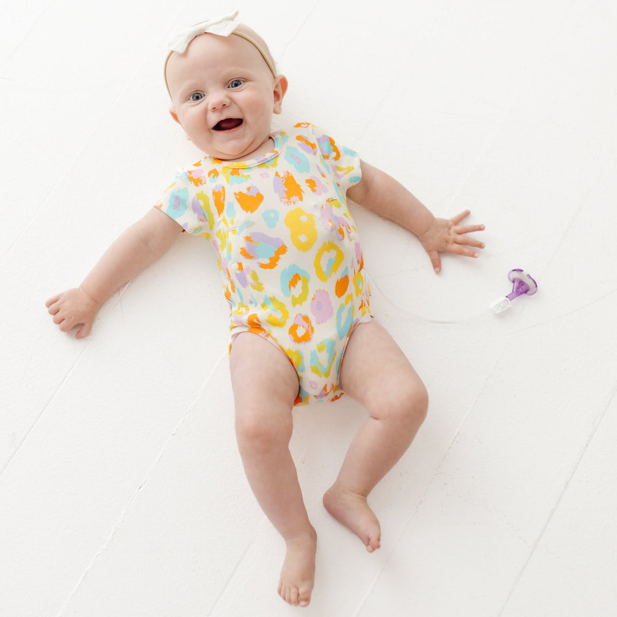 Baby wearing a colorful animal print onesie on a white floor.
