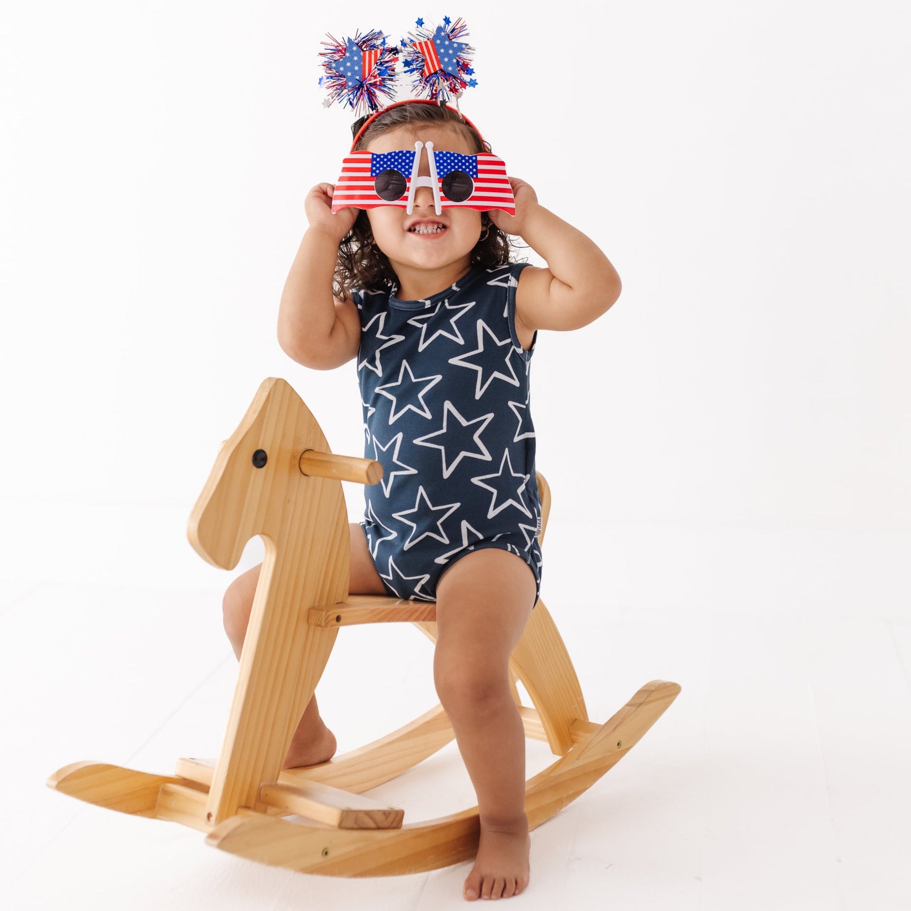 Child sitting on a wooden rocking horse wearing a blue star swimsuit and red, white, and blue headbands.