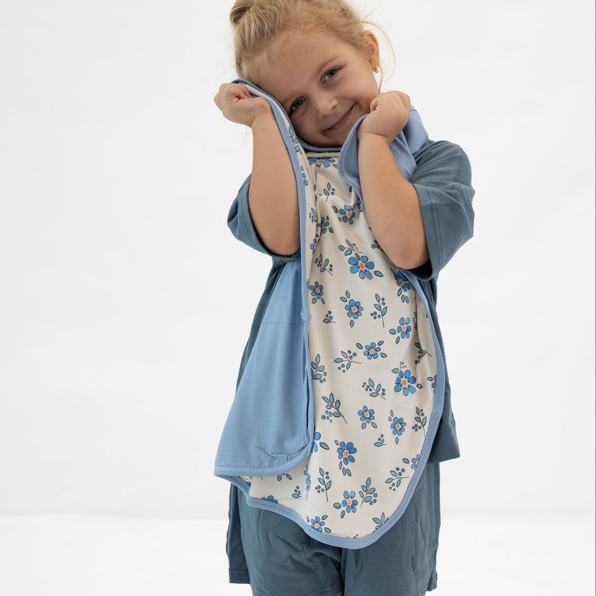 A young child wearing a blue short-sleeve shirt and shorts, holding up a reversible bib. The bib has a light blue outer layer and a cream-colored inner layer with a blue floral pattern, shown against a plain white background.