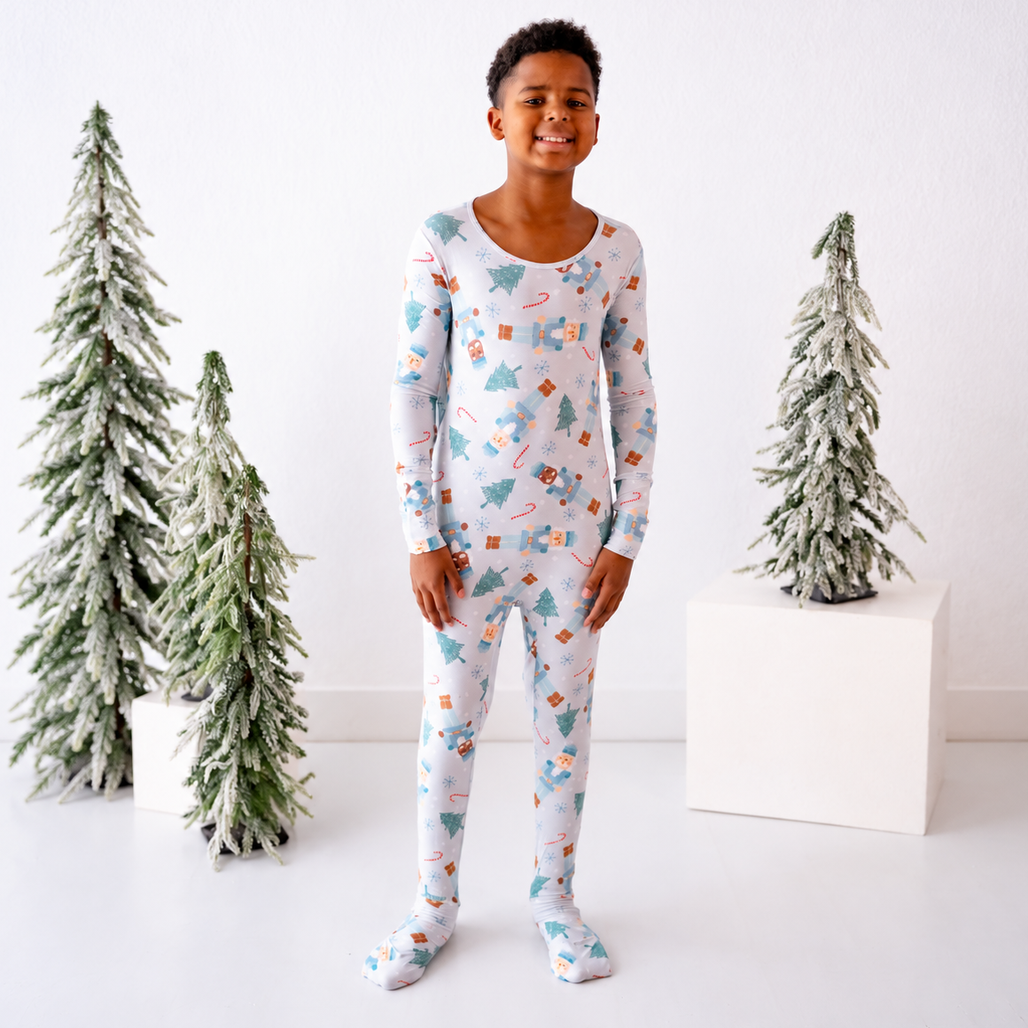 A young boy stands in a bright white room wearing a long-sleeve, full-length pajama set with a Christmas-themed print of trees, candy canes, and ornaments. Two small flocked Christmas trees are displayed on white pedestals on either side of him.
