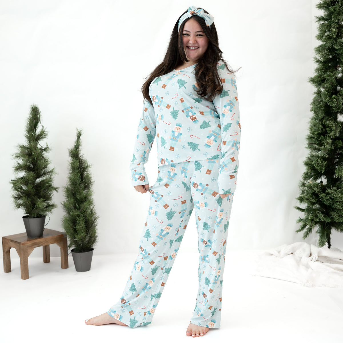 A woman modeling a light blue long-sleeve pajama set with a Christmas print, including a matching headband, standing in a studio decorated with artificial Christmas trees.