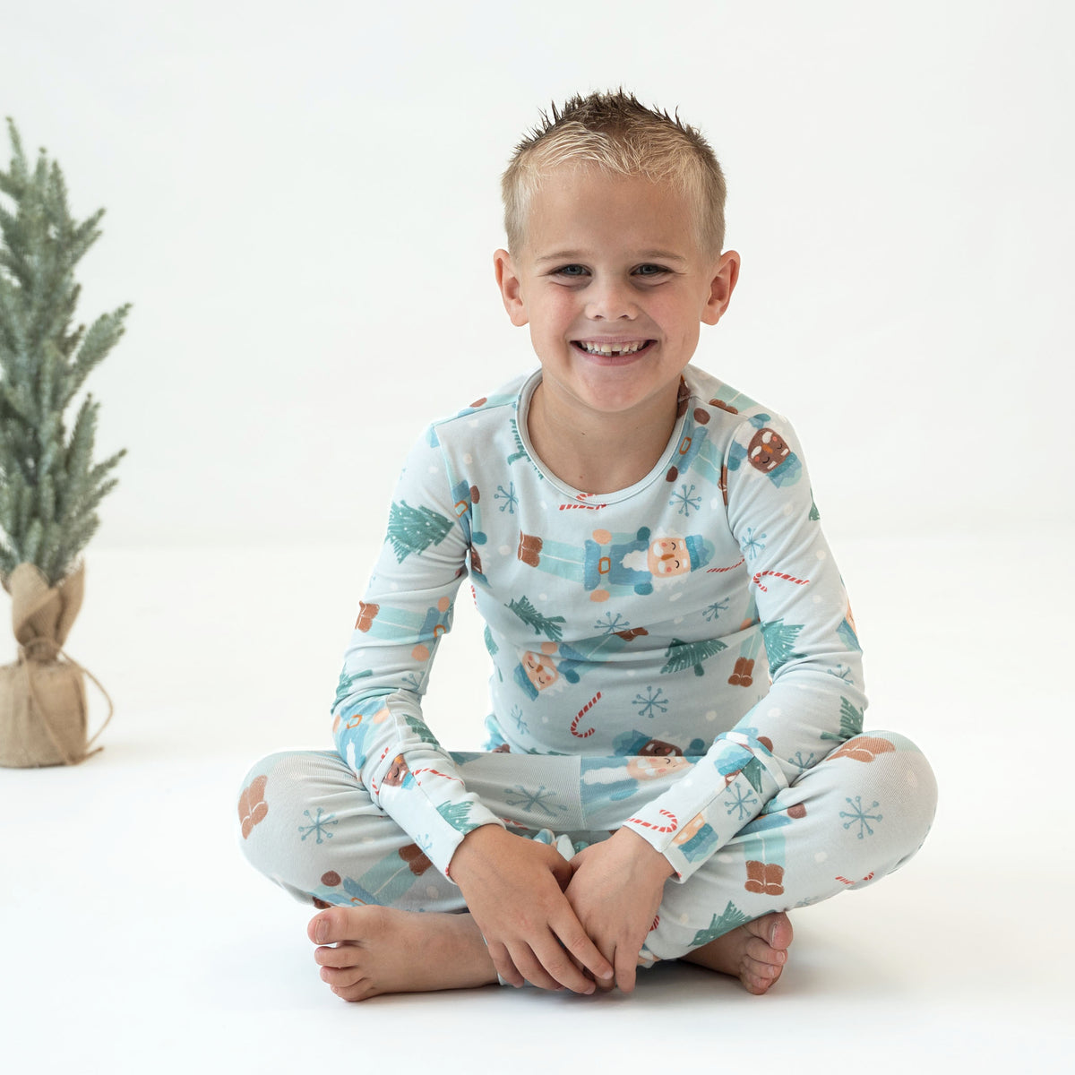 A young boy sitting cross-legged on a white studio floor, wearing a long-sleeve pajama top and matching pants with a light blue holiday print featuring gingerbread cookies, candy canes, snowflakes, and trees. A small potted pine tree is visible on the left side of the background.