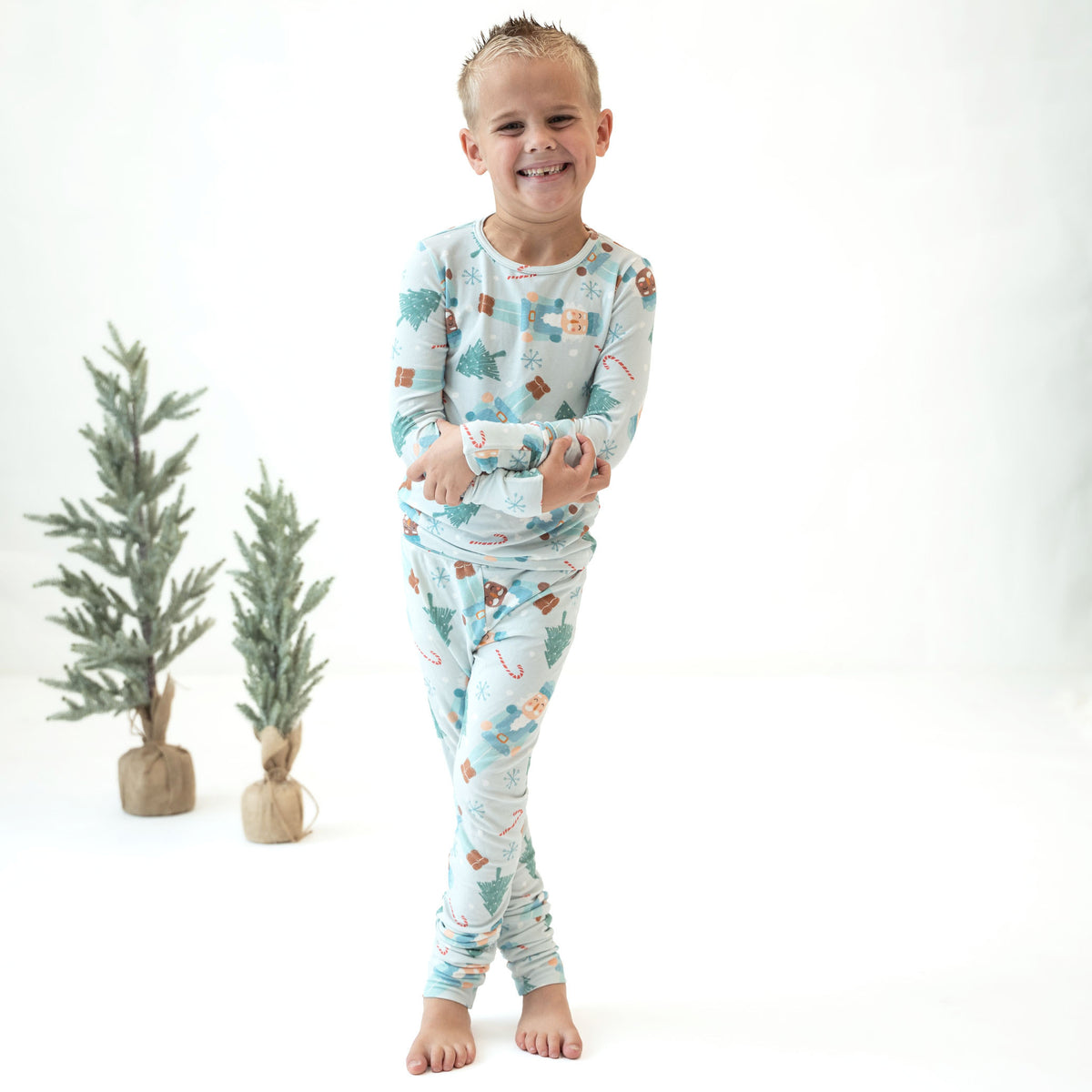 A smiling young boy stands barefoot in a studio wearing a light blue long-sleeve pajama set with a Christmas tree and snowman print. Two small potted trees sit to the left against a clean white background.