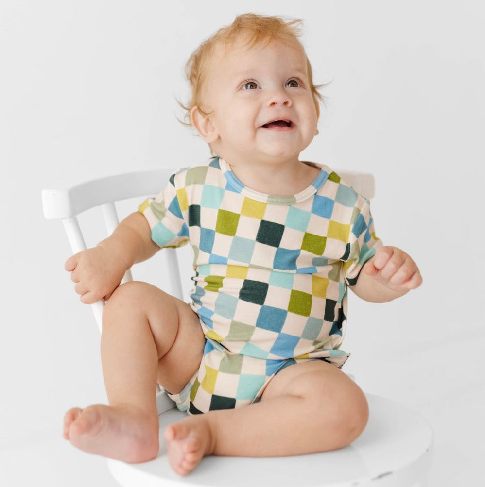 Baby wearing a colorful checkered onesie sitting on a white chair.