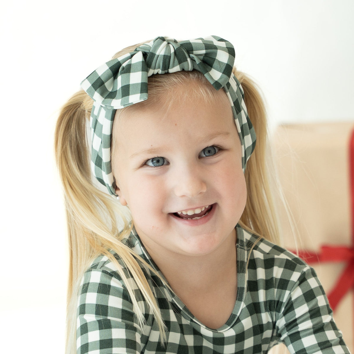 A young girl with blonde pigtails smiling, wearing a green and white gingham bow headband and a matching green and white gingham long-sleeve top. A wrapped gift box with a red ribbon is blurred in the background against a white studio backdrop.