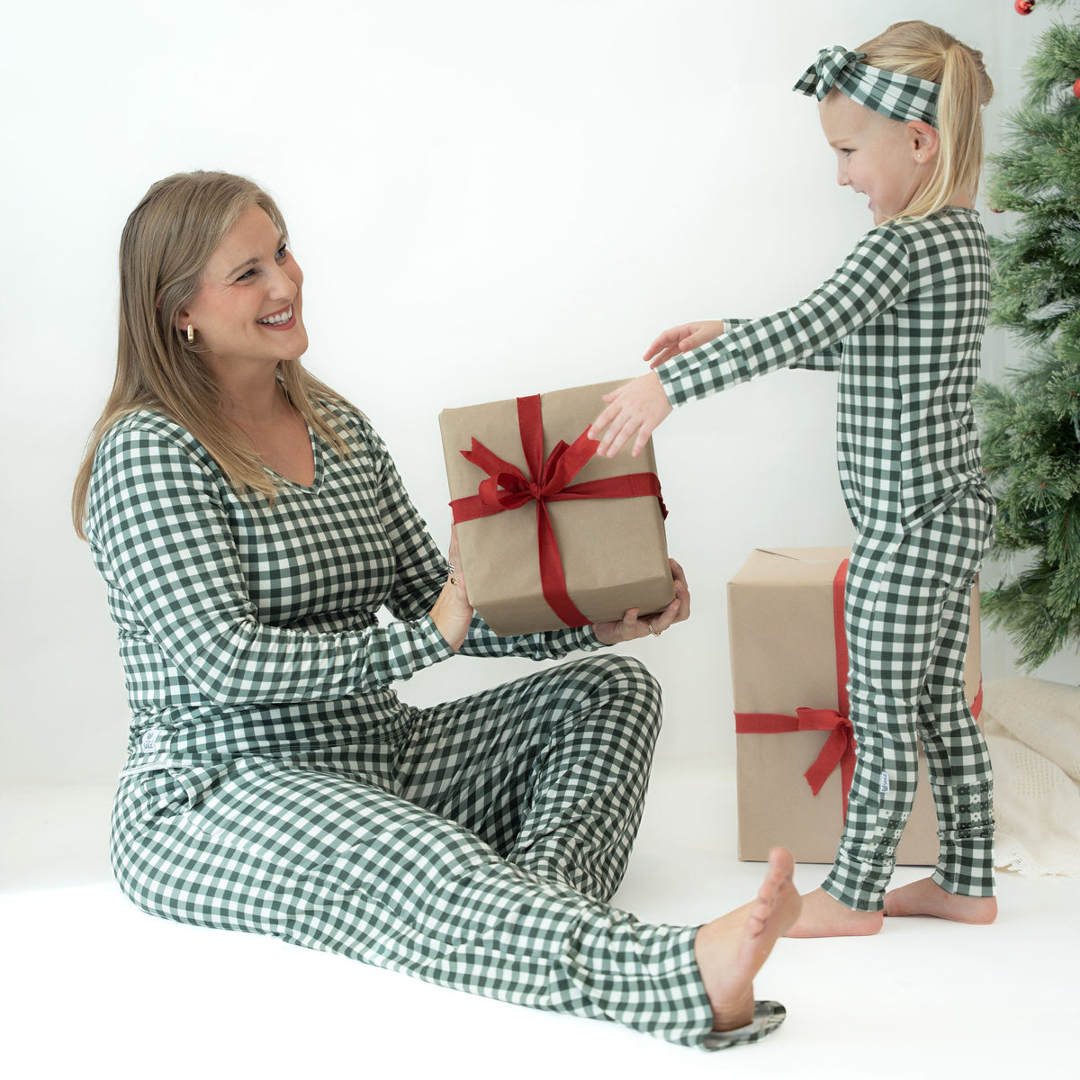 A woman and a young girl wearing matching green and white gingham pajamas share a wrapped gift box in a bright white room with a Christmas tree in the background.